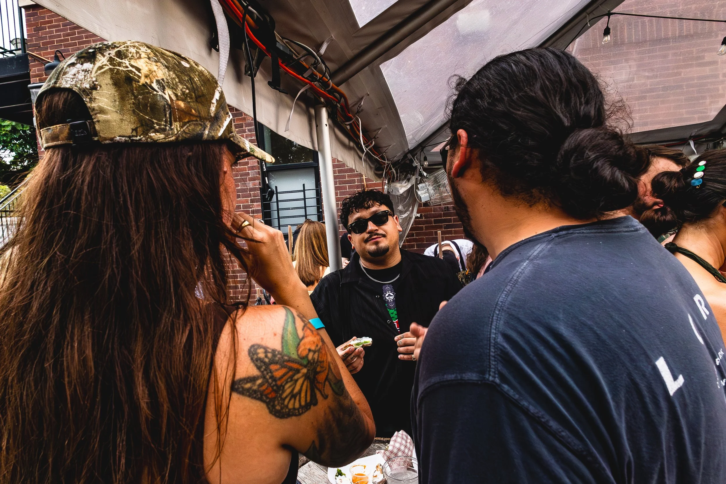 Group of people talking at an outdoor event under a canopy, with brick building in the background. One woman with long hair and a butterfly tattoo on her arm is wearing a camouflage hat, and a man with dark hair and sunglasses is holding a snack whil