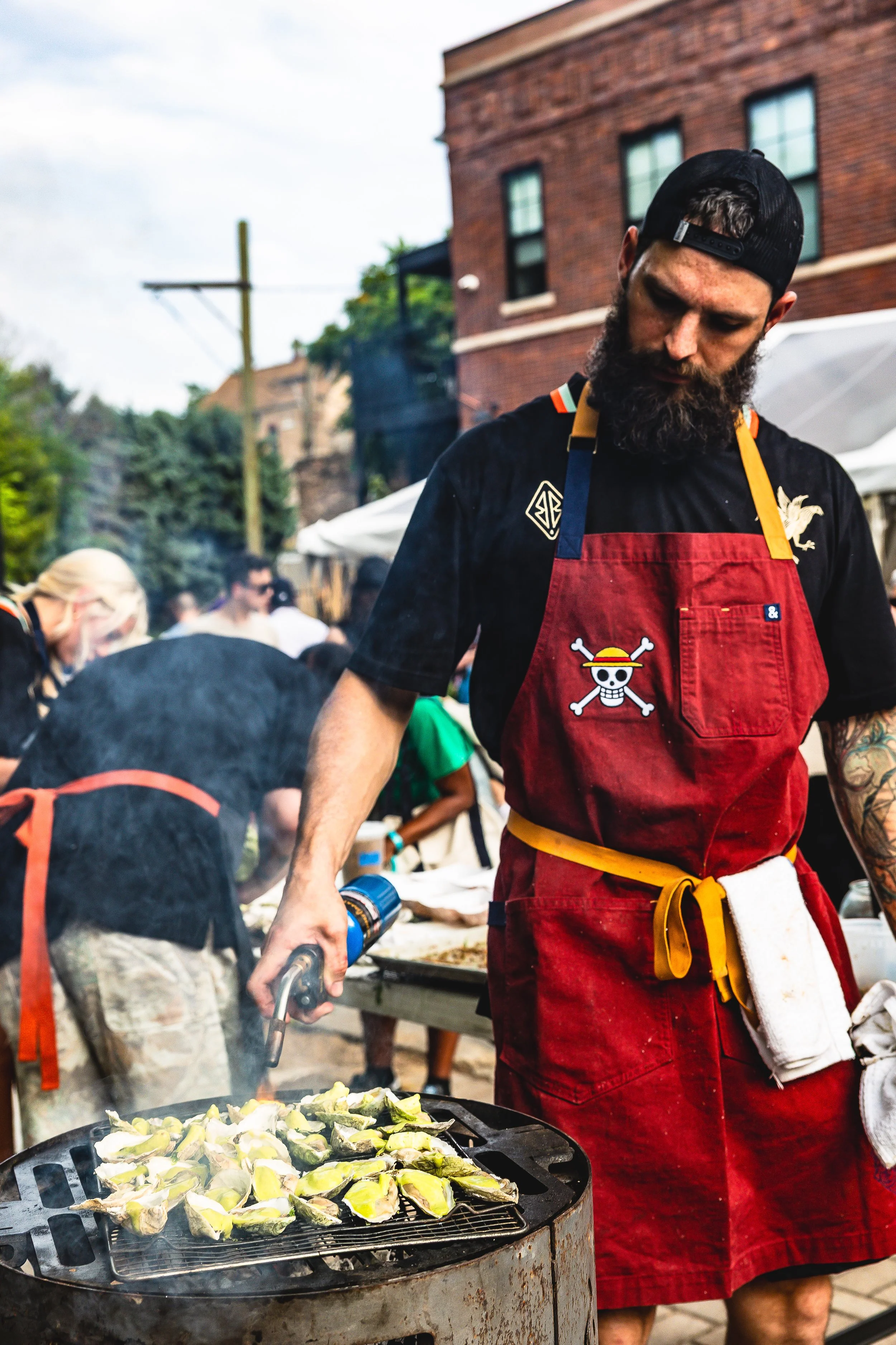 A man with a beard and black cap grilling vegetables outdoors at a street event, wearing a red apron with a skull pirate logo.