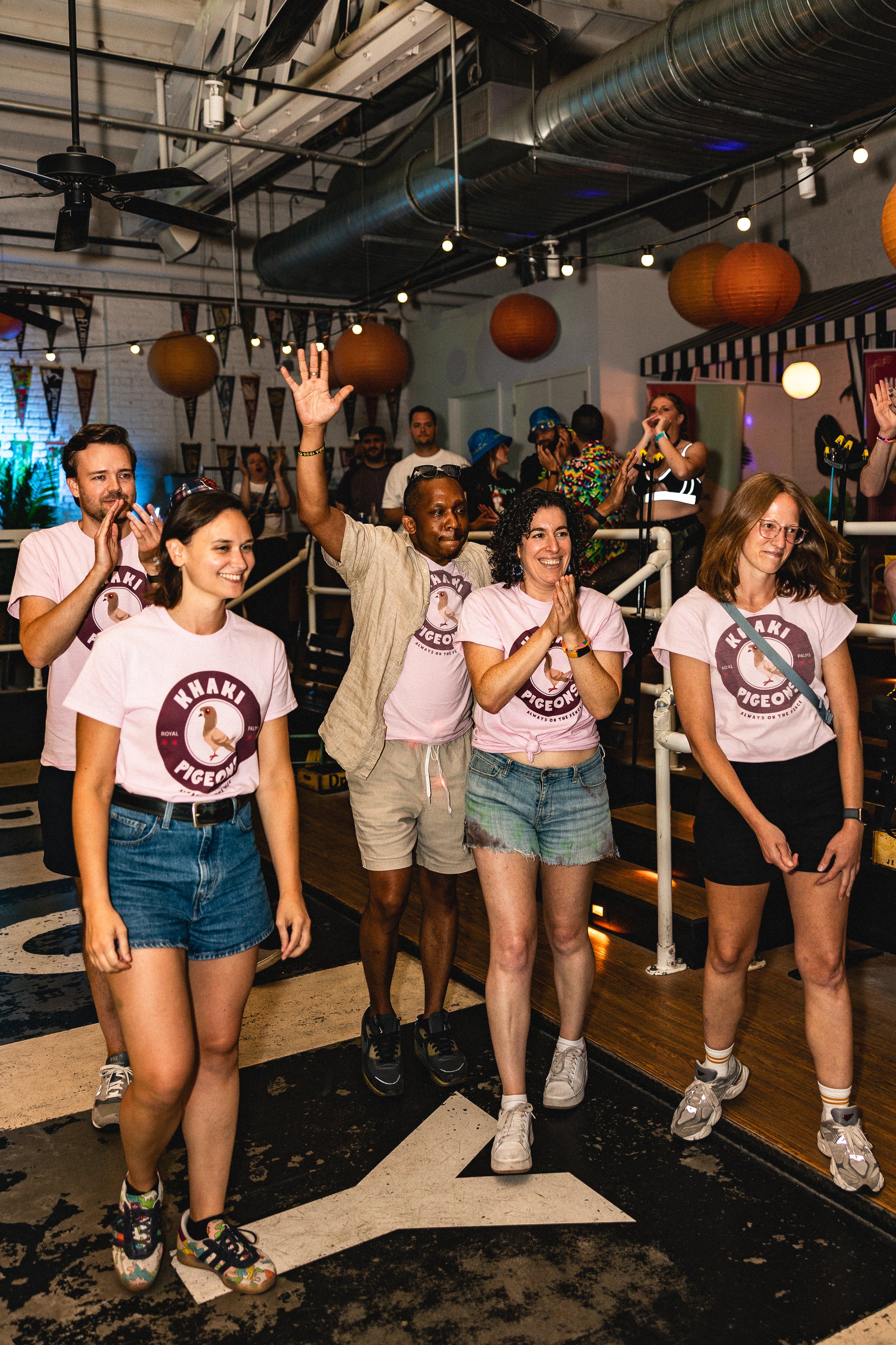 Group of people at a party, smiling and clapping, some wearing matching pink t-shirts with a pigeon logo, inside a decorated indoor venue.