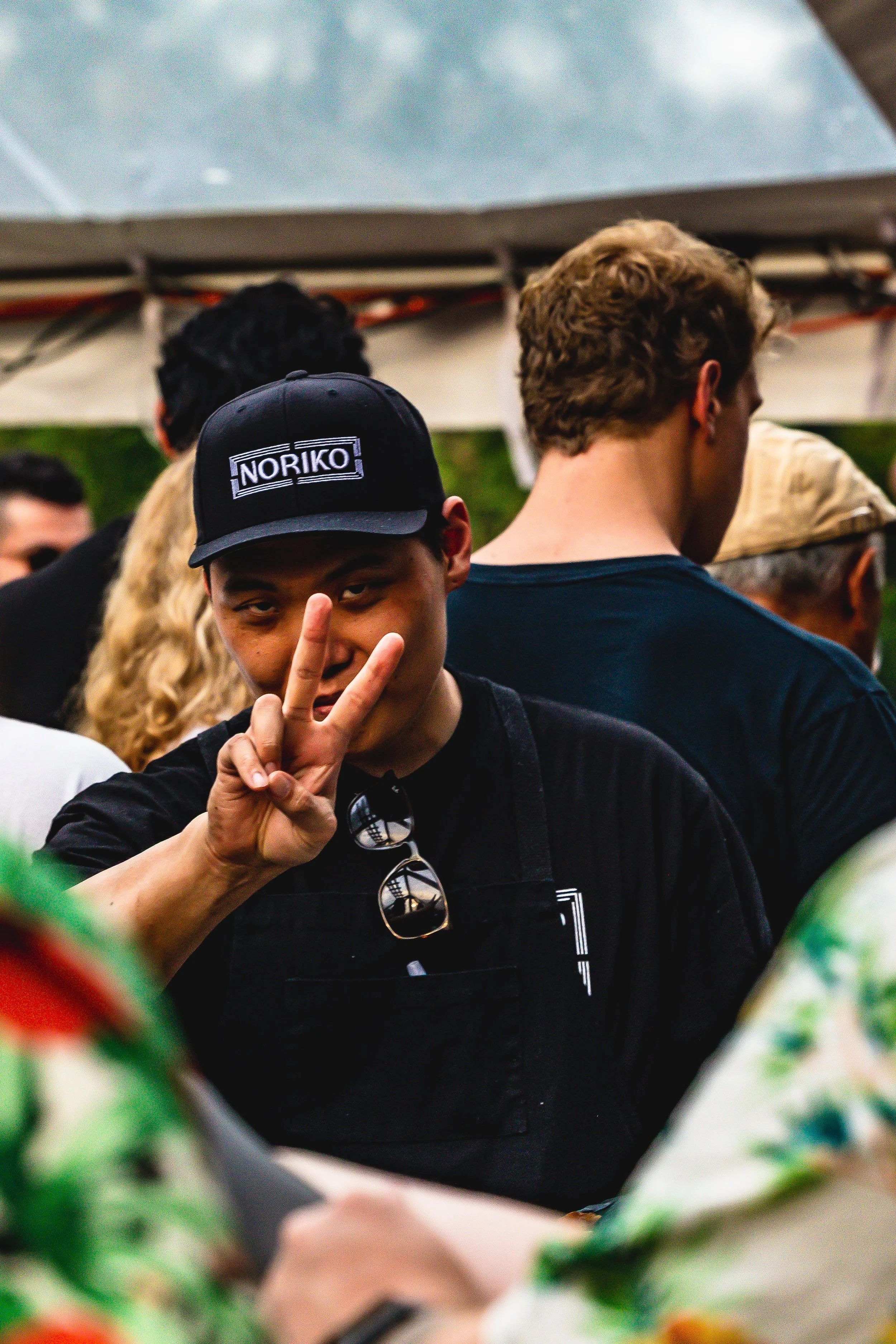 A young man with a black cap that reads Noriko, making a peace sign and smiling slightly at the camera at an outdoor gathering.