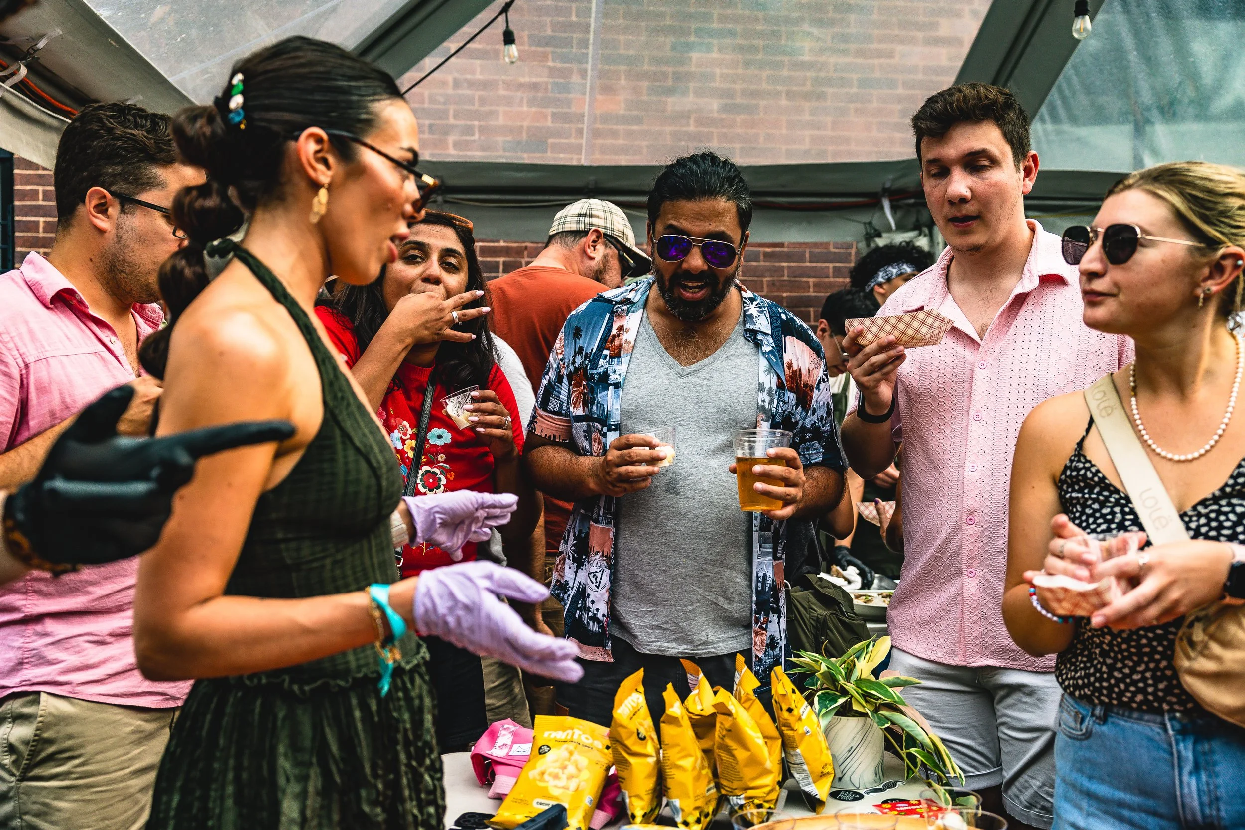 A group of people socializing at a backyard gathering, with snacks, drinks, and a brick wall in the background.