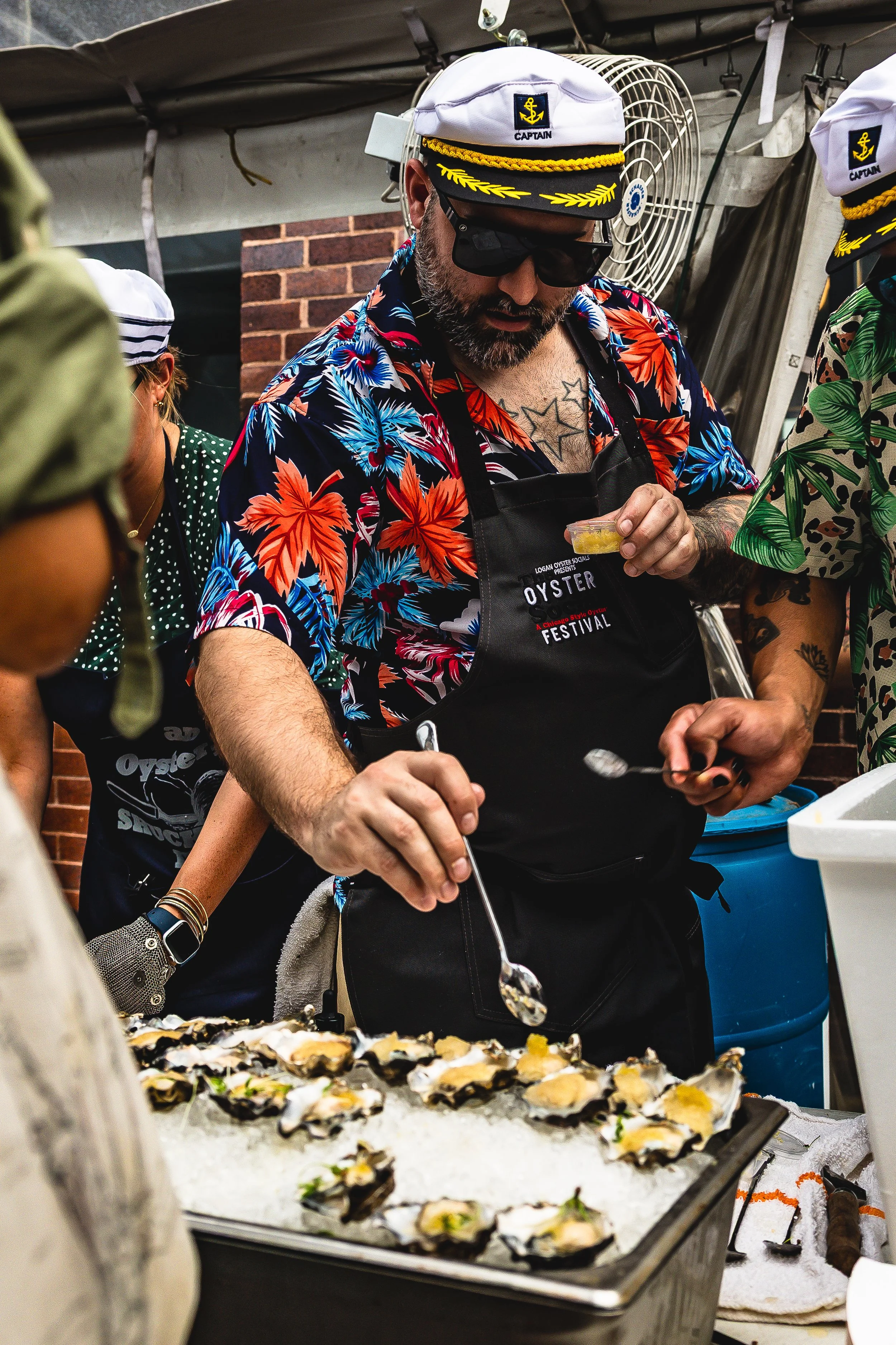 A man wearing a colorful Hawaiian shirt, a captain's hat, sunglasses, and an apron serving oysters on a bed of ice at a seafood festival.