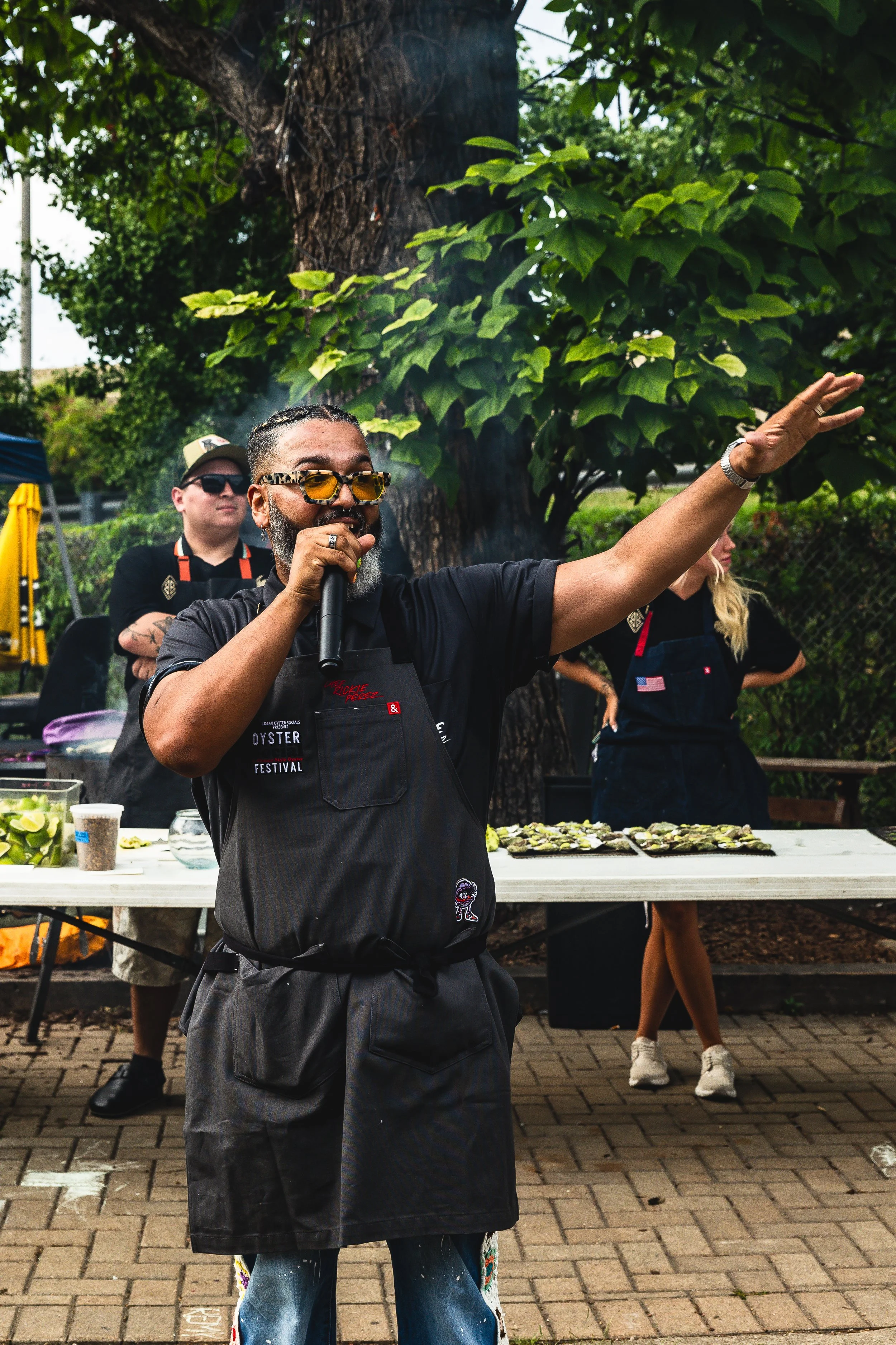 A man with sunglasses and a beard speaking into a microphone at an outdoor event, with a table of oysters behind him, and trees in the background.