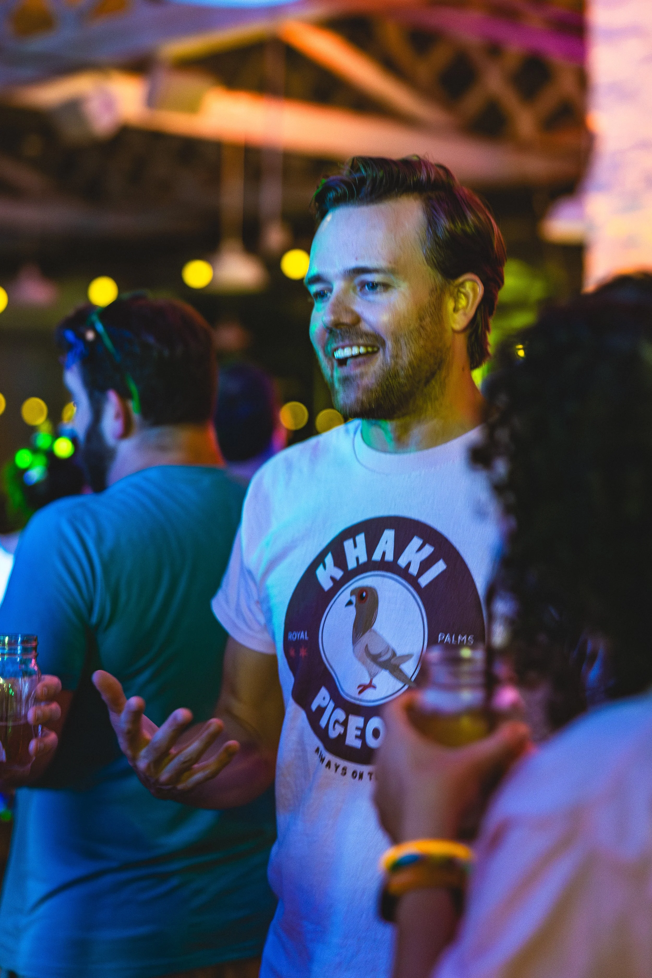 A man with a beard and styled hair smiling and talking at a party or social gathering, wearing a white t-shirt with a graphic of a bird and the words "Kha Ki Pigeo" on it, holding a drink in his hand.
