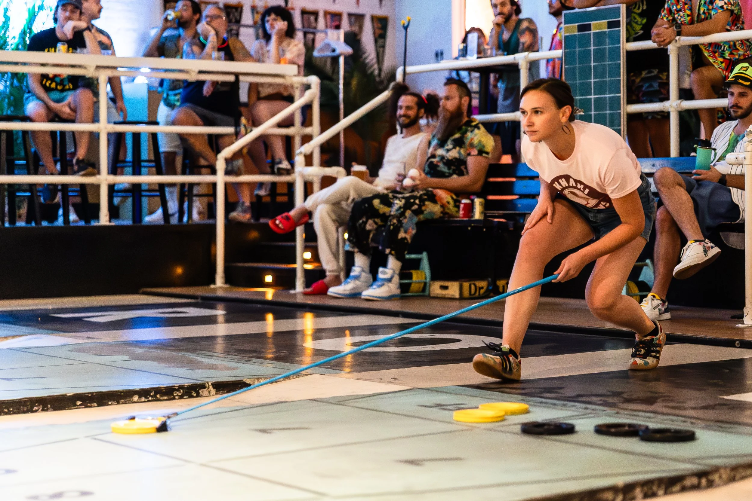A woman playing shuffleboard on an indoor court with several spectators watching from the stands and sitting around the court.