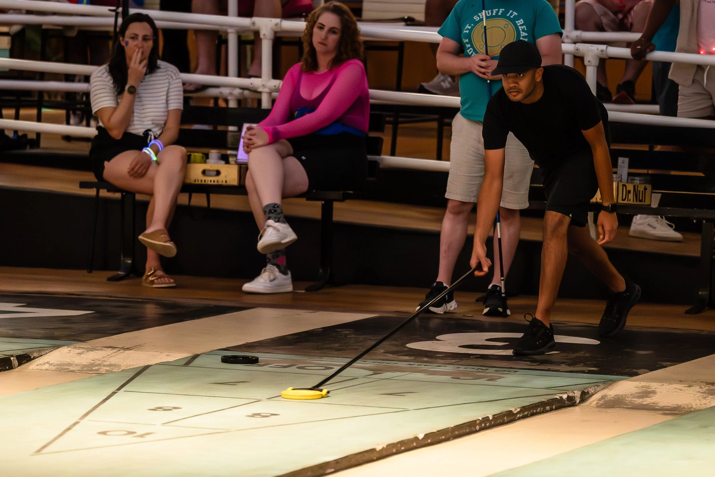 A man playing shuffleboard while three women sit on benches watching in an indoor game area.