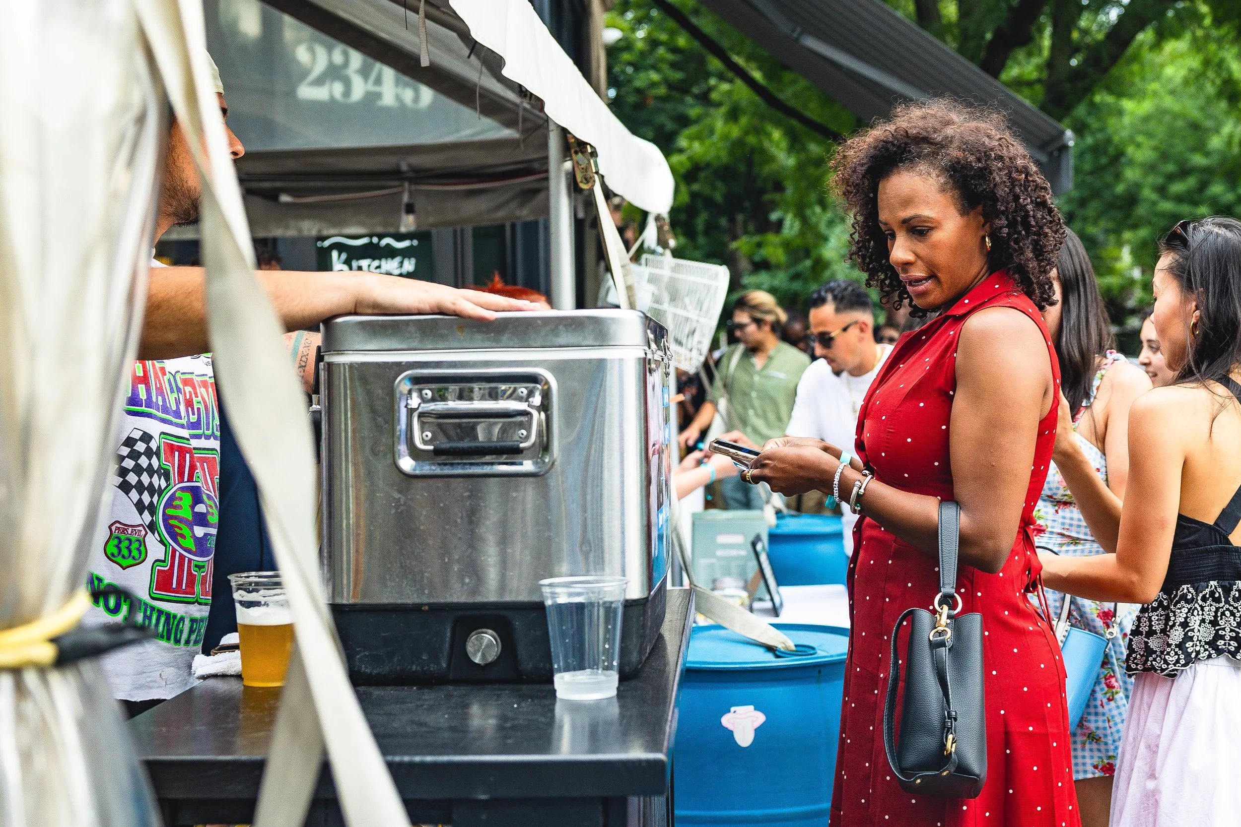 A woman in a red dress with white polka dots looks at her phone while standing at a food stall at an outdoor event, with several people in the background.