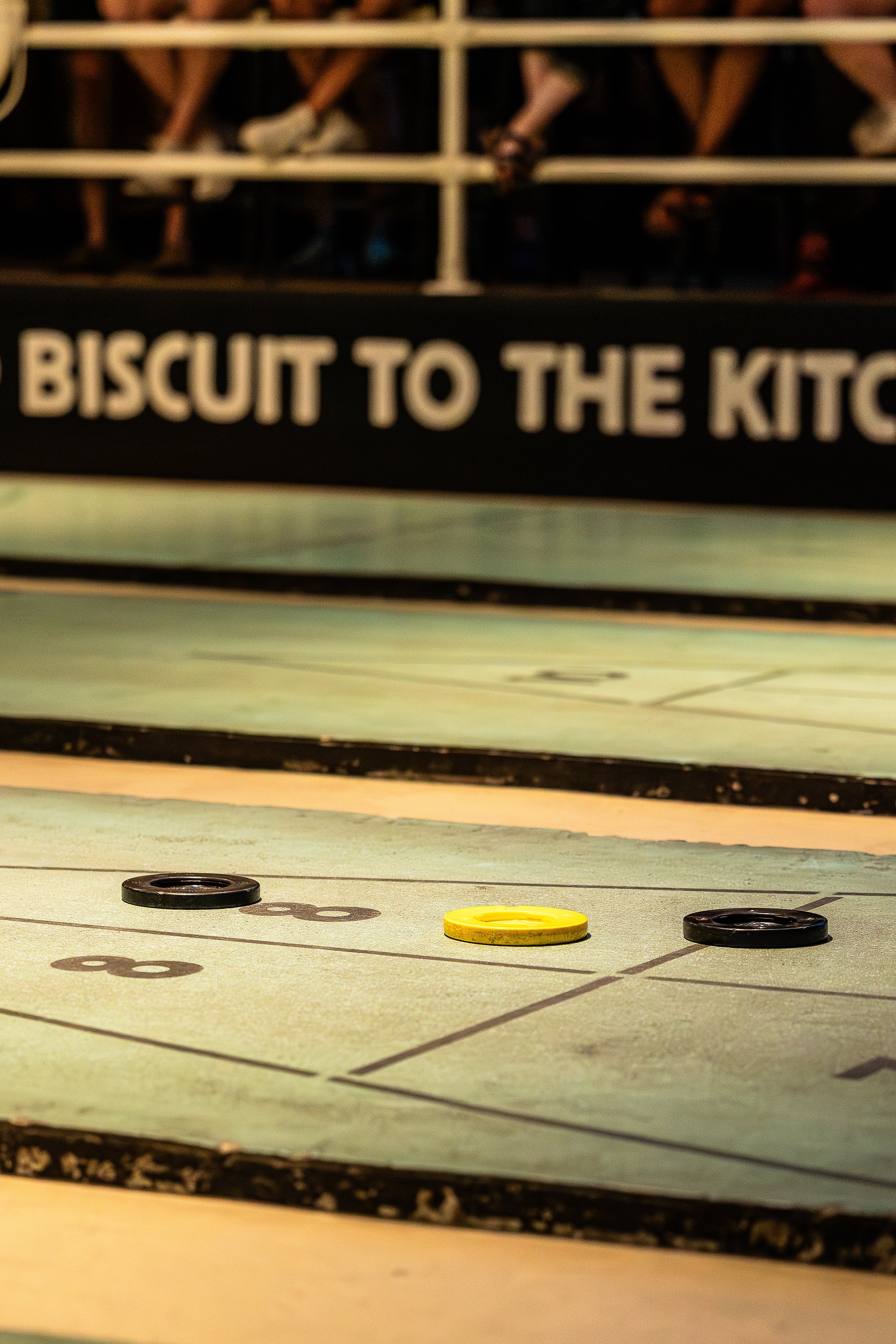 Close-up view of a shuffleboard court with colored pucks, with a sign in the background that reads "BISCUIT TO THE KITCHEN."