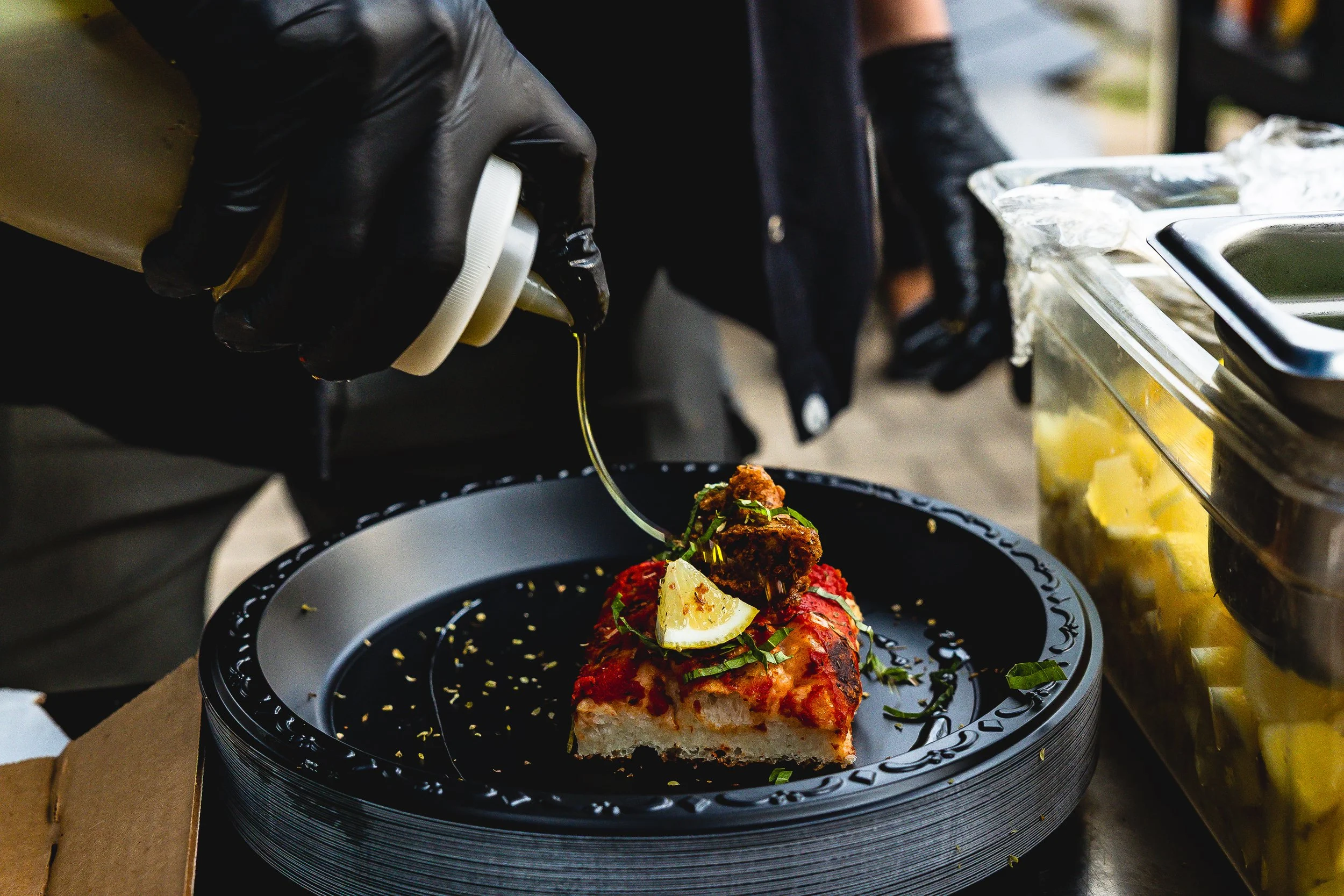 Person in black gloves pouring sauce on a slice of pizza with toppings, lemon wedge, and herbs