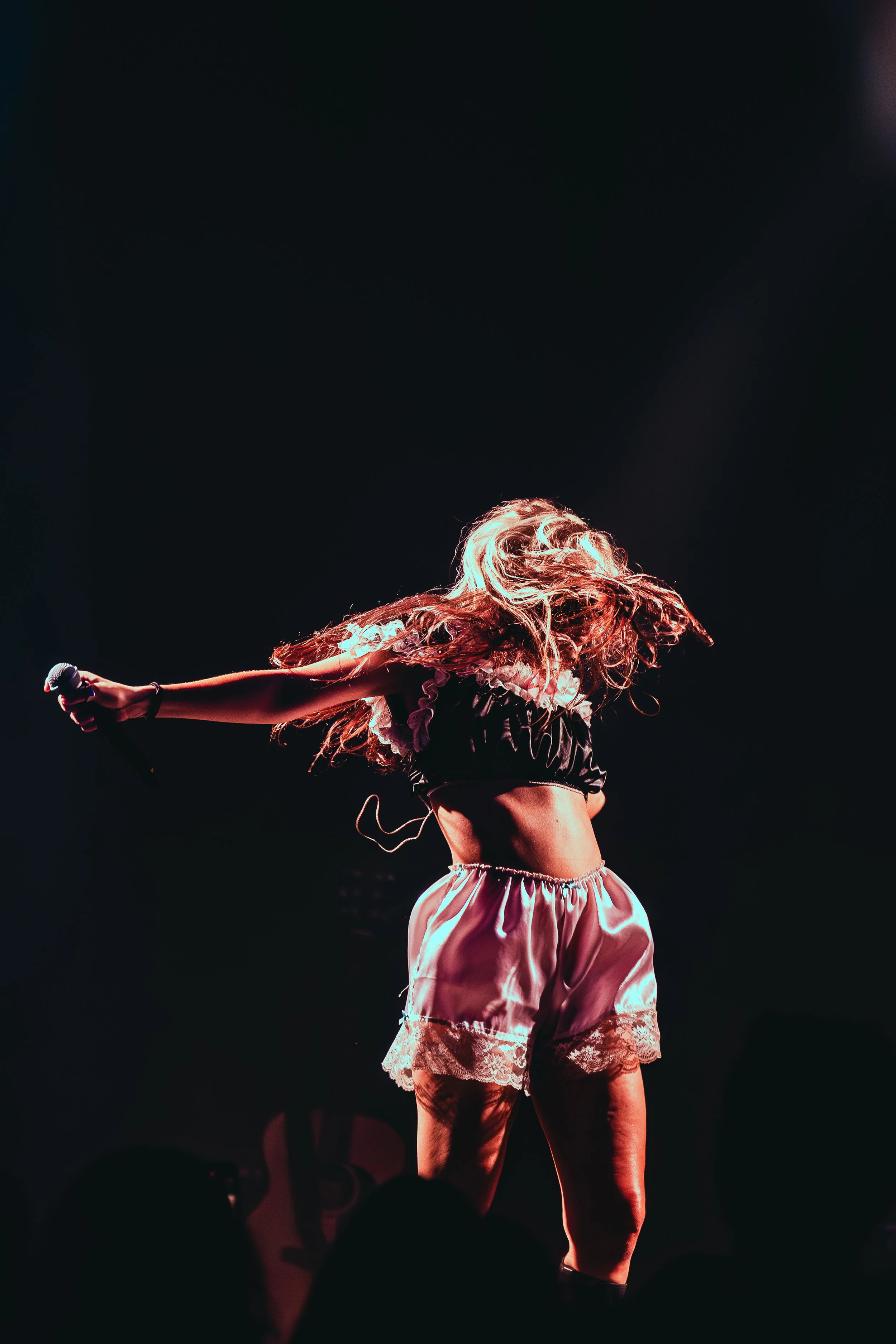 A female performer on stage with long hair, wearing a black crop top and satin shorts with lace trim, holding a microphone and dancing.