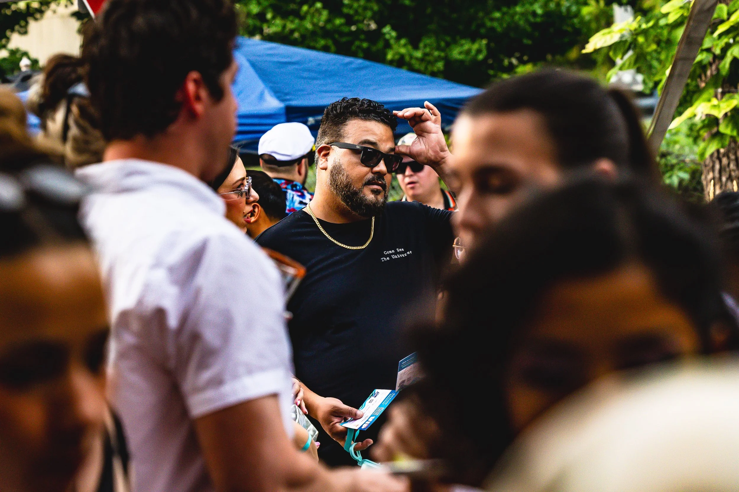 Man with sunglasses, gold chain, and black T-shirt among a crowd at an outdoor event, with blue tent and green trees in the background.