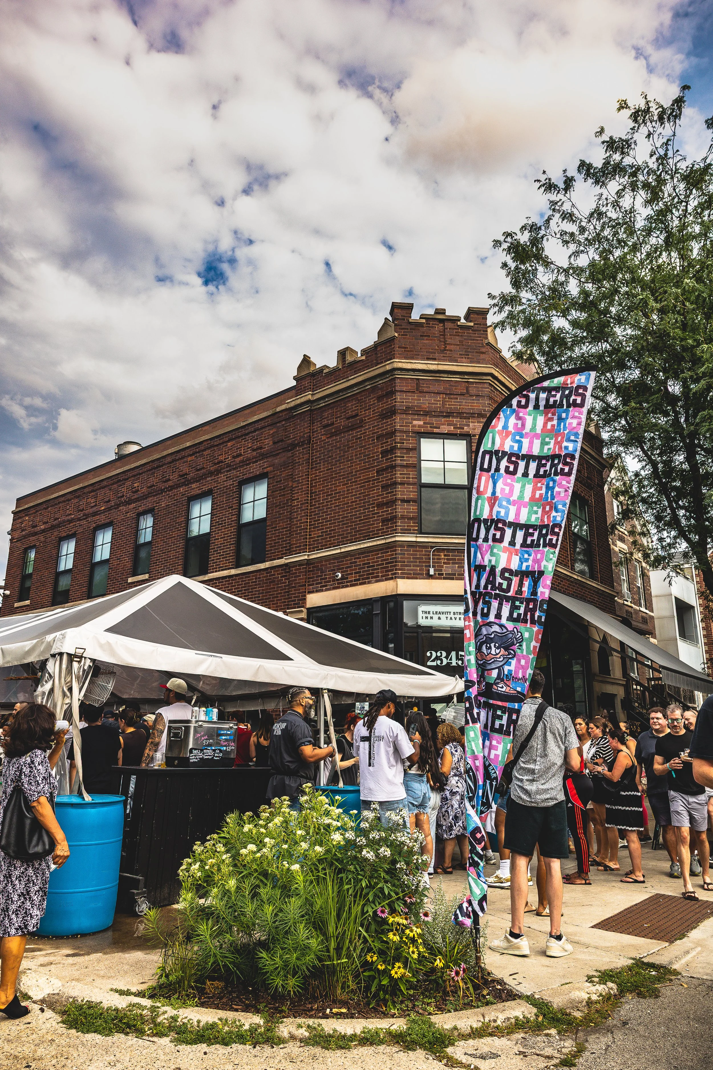 Crowd gathered outside a street food vendor under a white tent with a banner that reads 'Oysters' in colorful letters. The scene is set on a city street with a brick building in the background and a partly cloudy sky overhead.