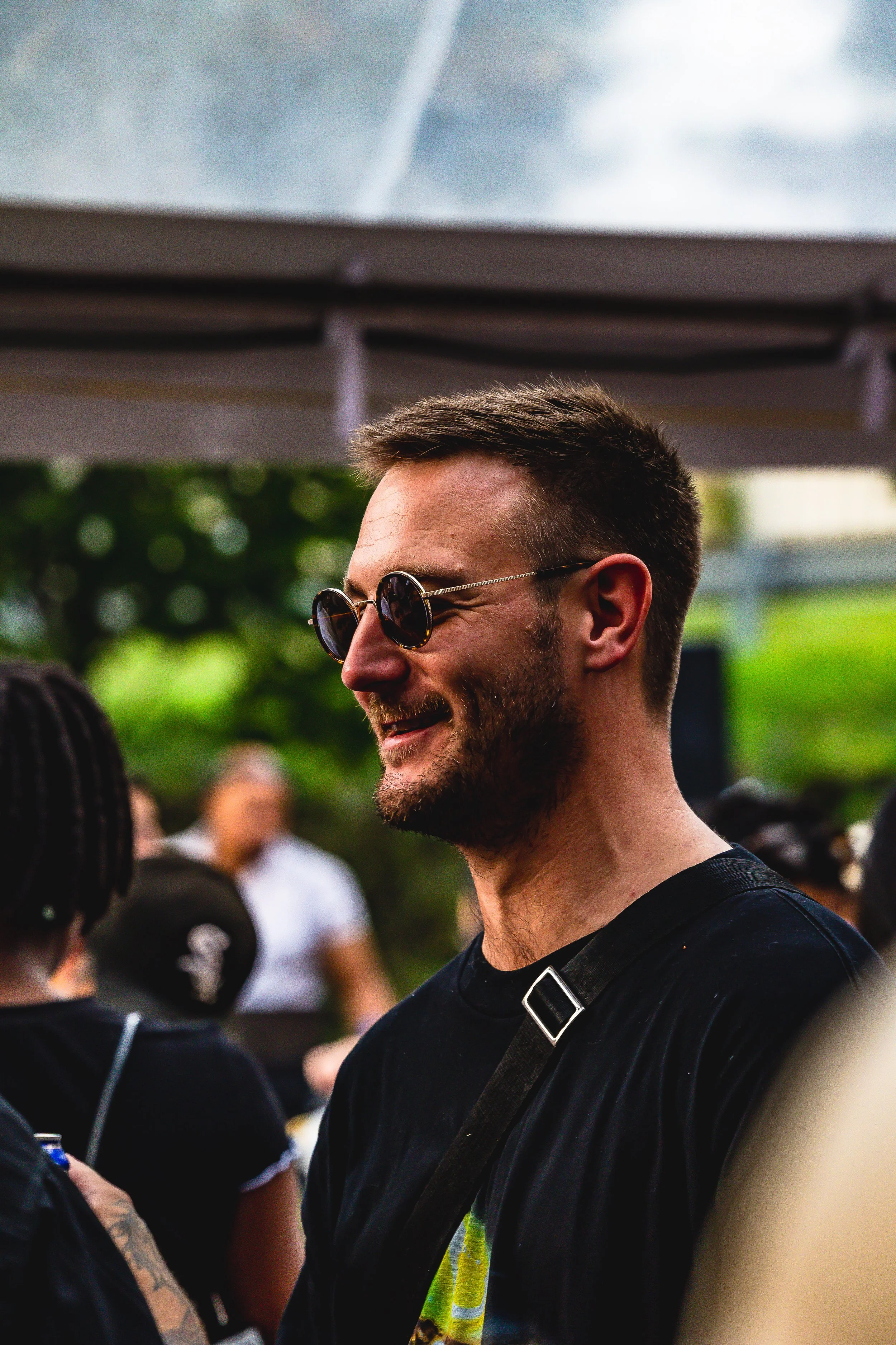 A man with short dark hair, a beard, and sunglasses smiling in a crowd outdoors.
