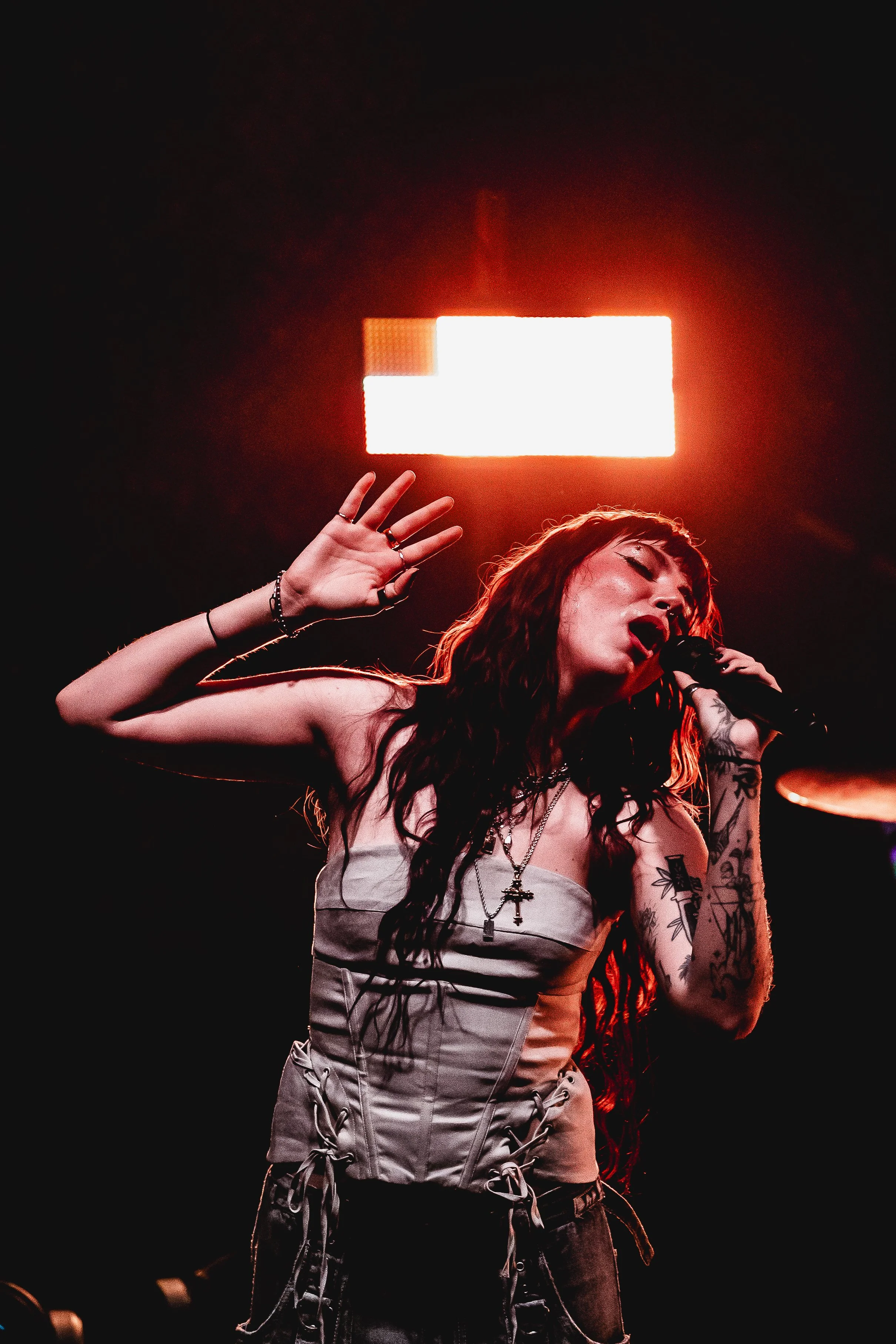 A woman singing into a microphone on stage with her eyes closed and one hand raised, illuminated by a bright red light in the background.