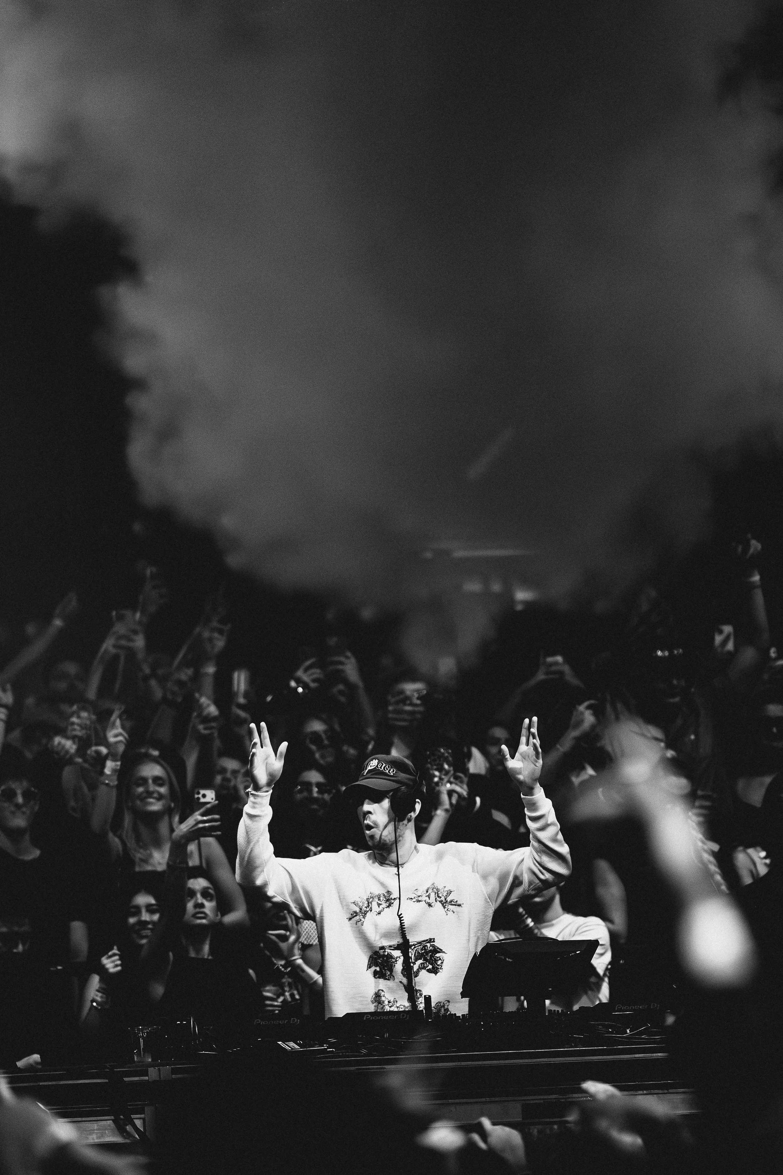Cloonee wearing headphones and a cap performs at a nightclub with a crowd dancing and taking photos.
