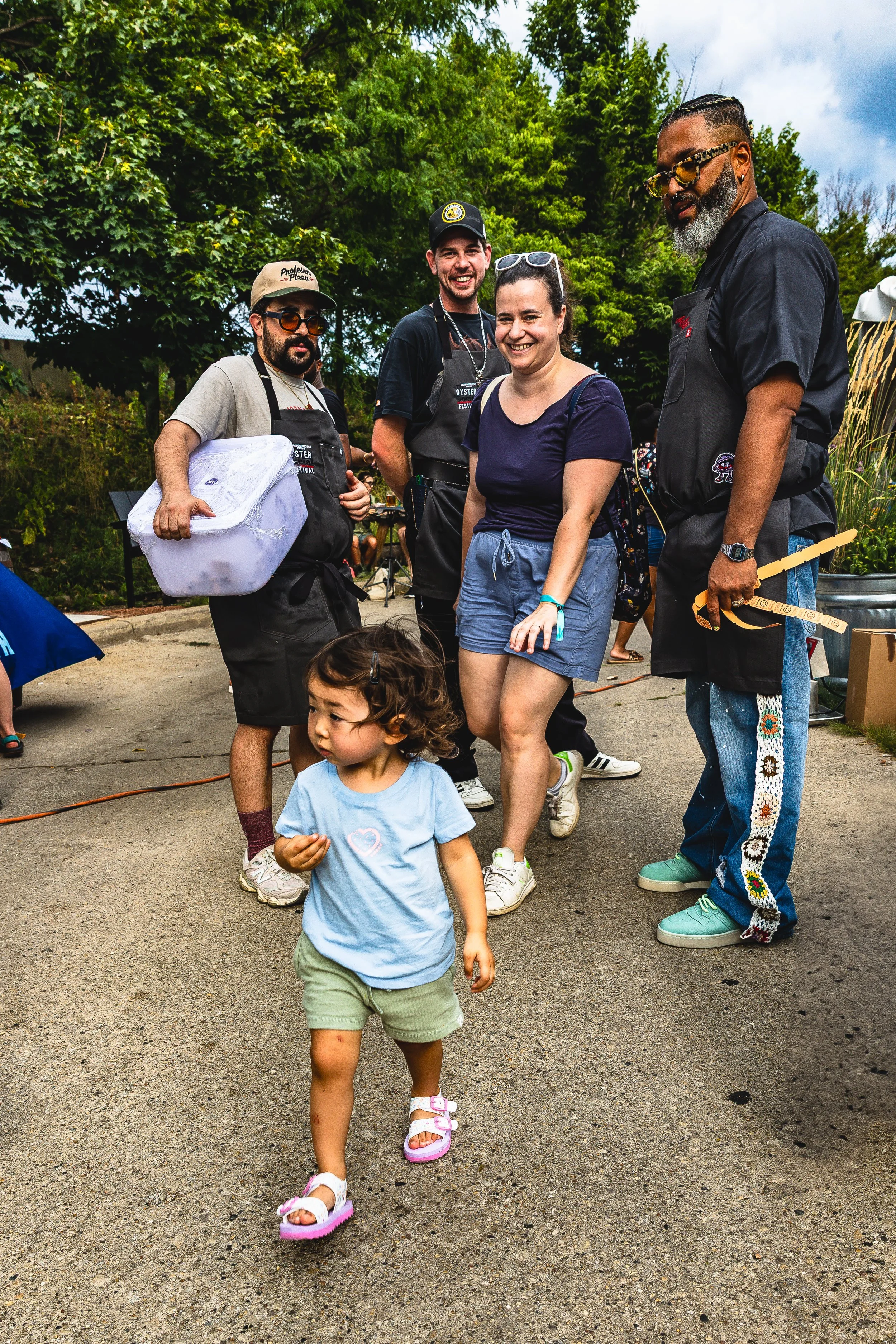 Group of people, including a child walking in front, outdoors with trees, enjoying a festival or gathering.