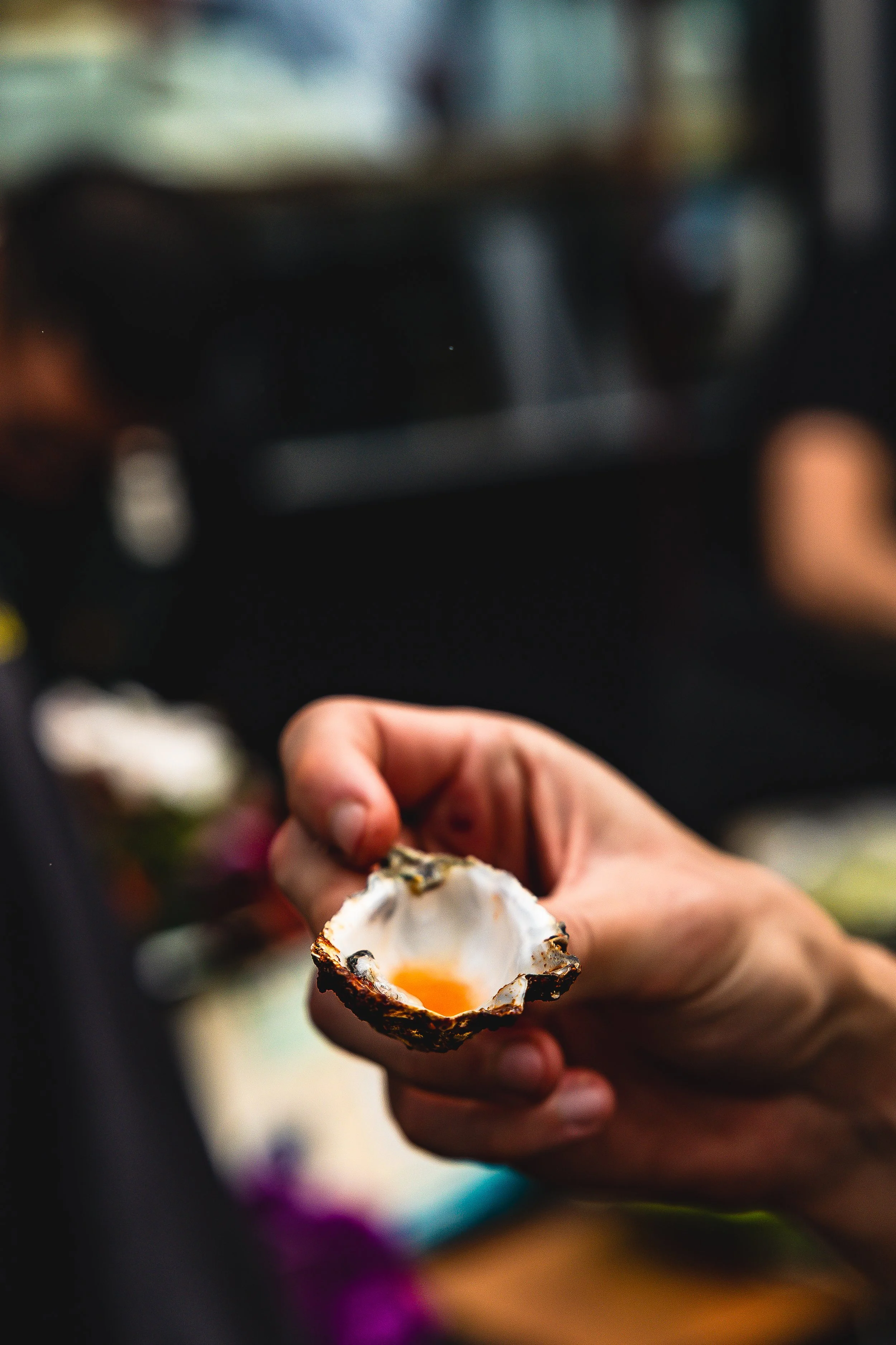 Close-up of a hand holding an empty oyster shell with a small amount of orange roe inside, against a blurred background.