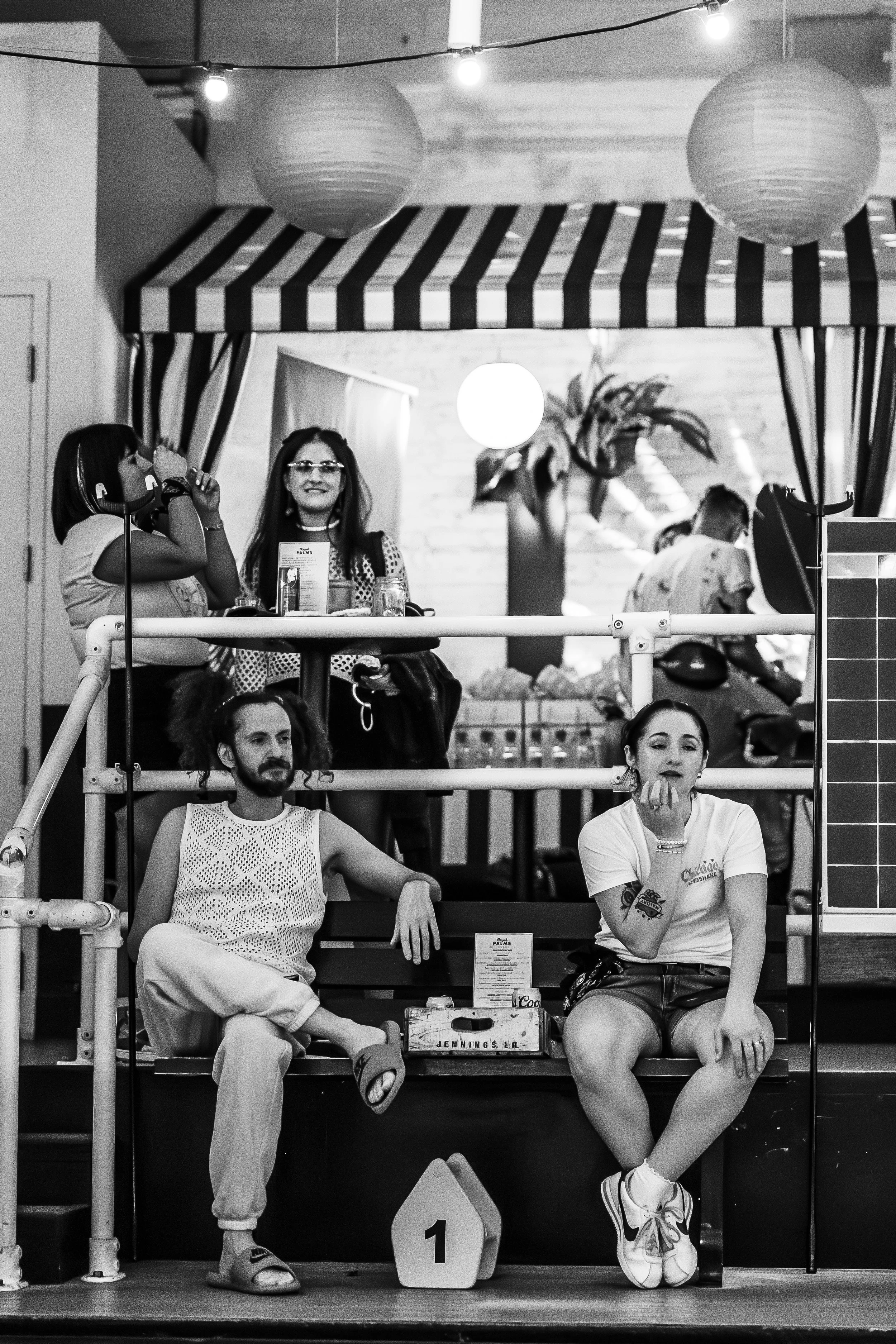A black-and-white photo of four people sitting and standing around an indoor space with a striped canopy, lanterns, and decorative plants. Some individuals are posing and smiling, while others are engaging in activities. There is a bench with a sign 