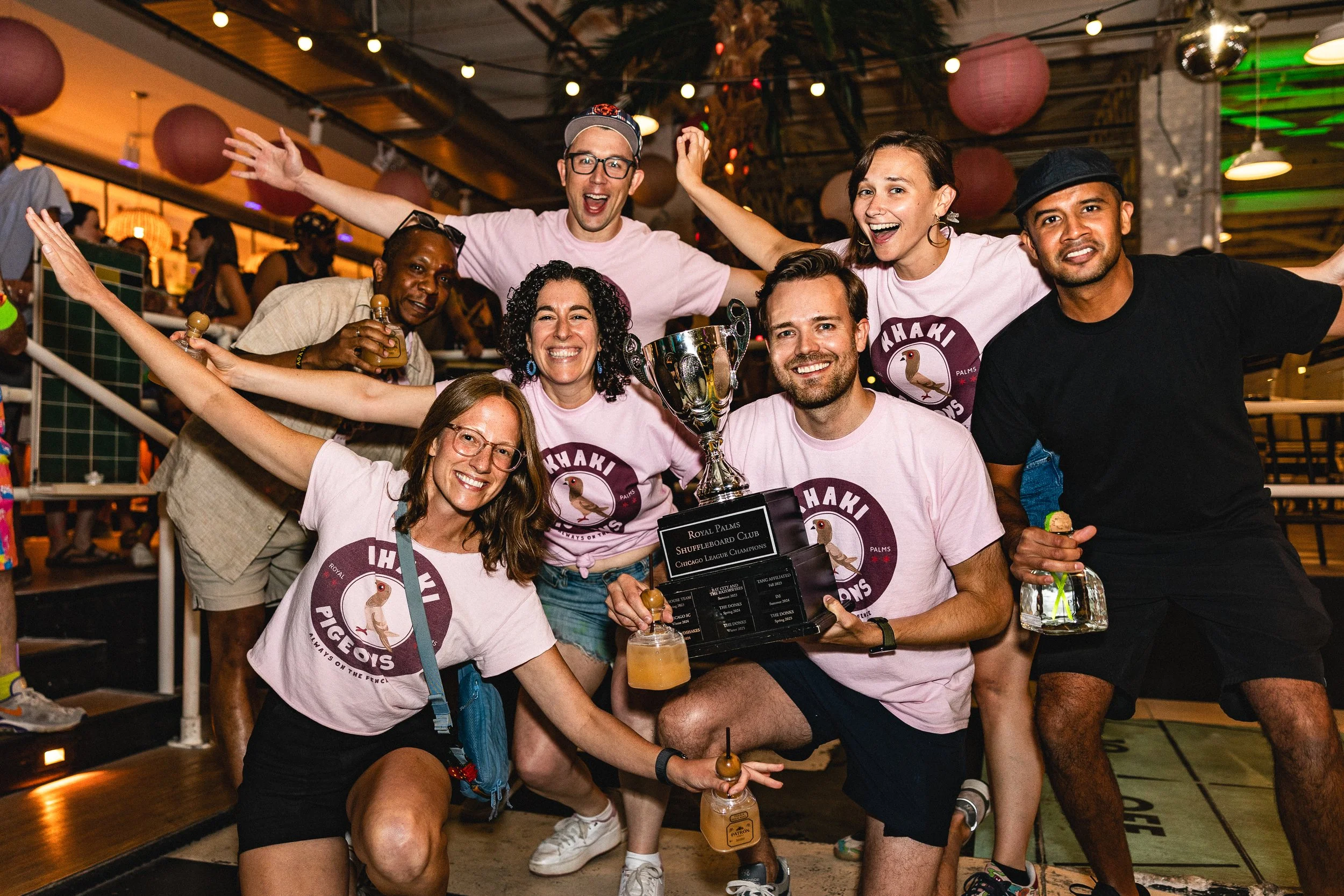 Group of people celebrating a victory at a shuffleboard club, holding a large trophy and drinks, smiling and posing for the camera.