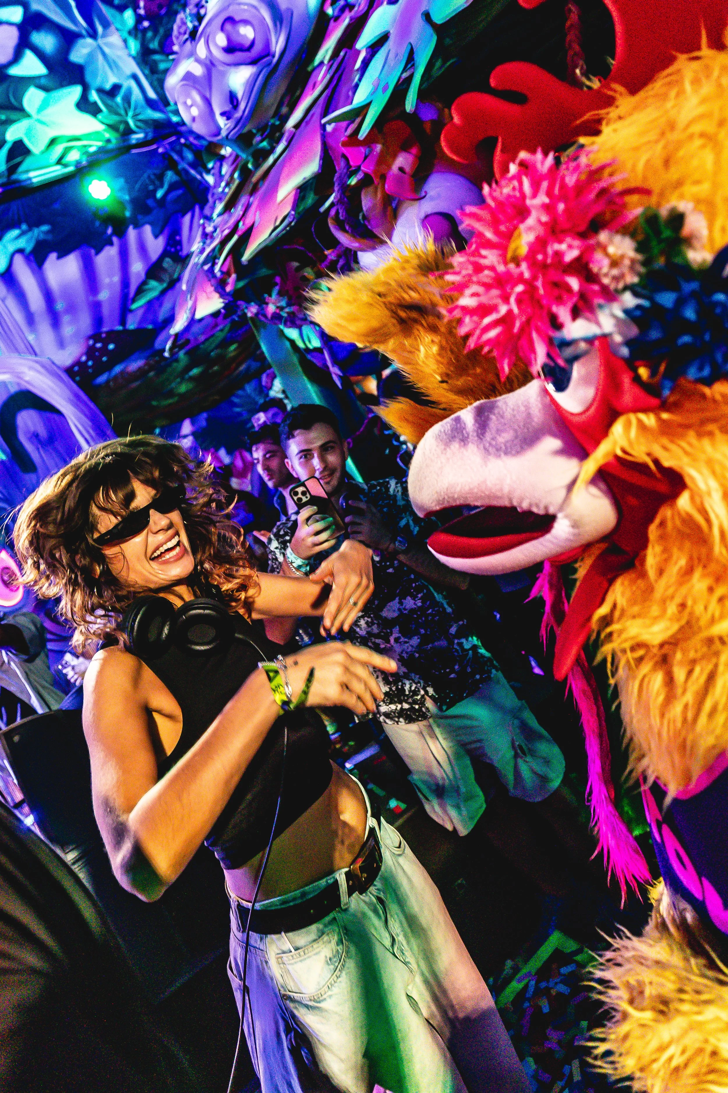 A woman dancing and having fun at a lively party with colorful lights, wearing black sunglasses and headphones around her neck, near a large puppet or mascot resembling a pink and orange bird with a beak and feathers.