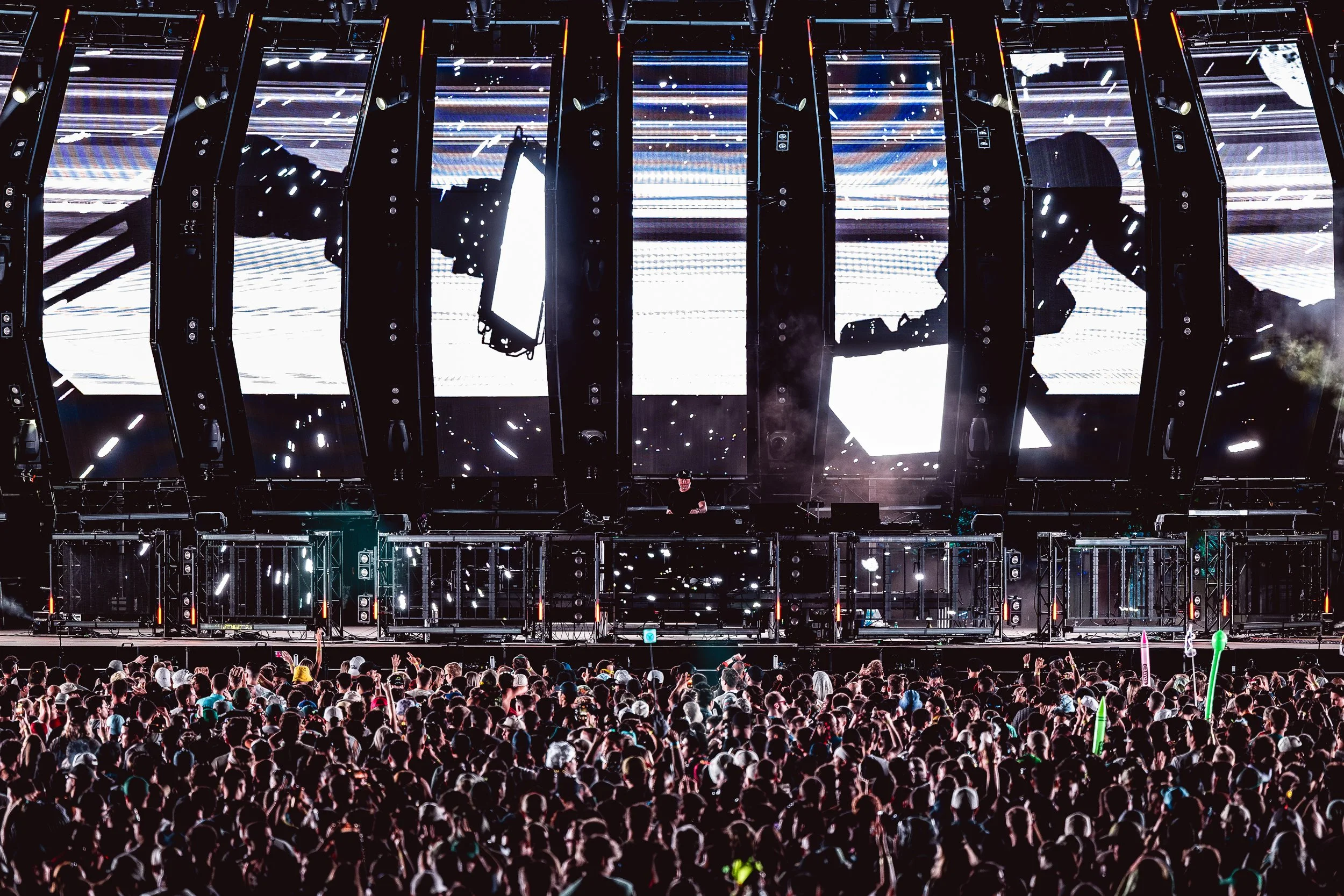 A large crowd of people at a concert with a DJ performing on stage illuminated by large digital screens and colorful lights.