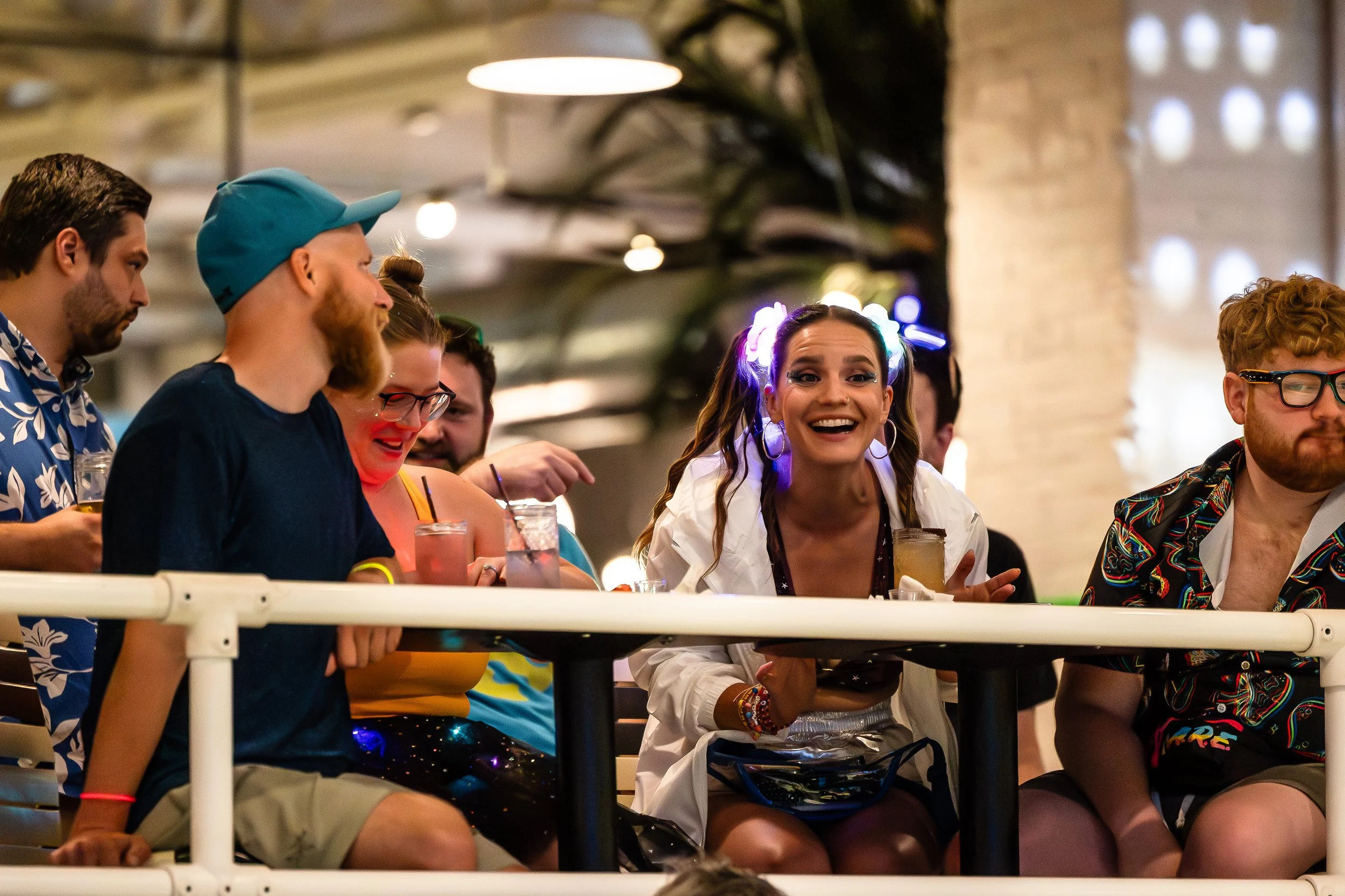 Group of young adults sitting at a bar or club, smiling and enjoying drinks, dressed in colorful, casual clothing, with some wearing glasses and hairbands, in a lively indoor setting with warm lighting.