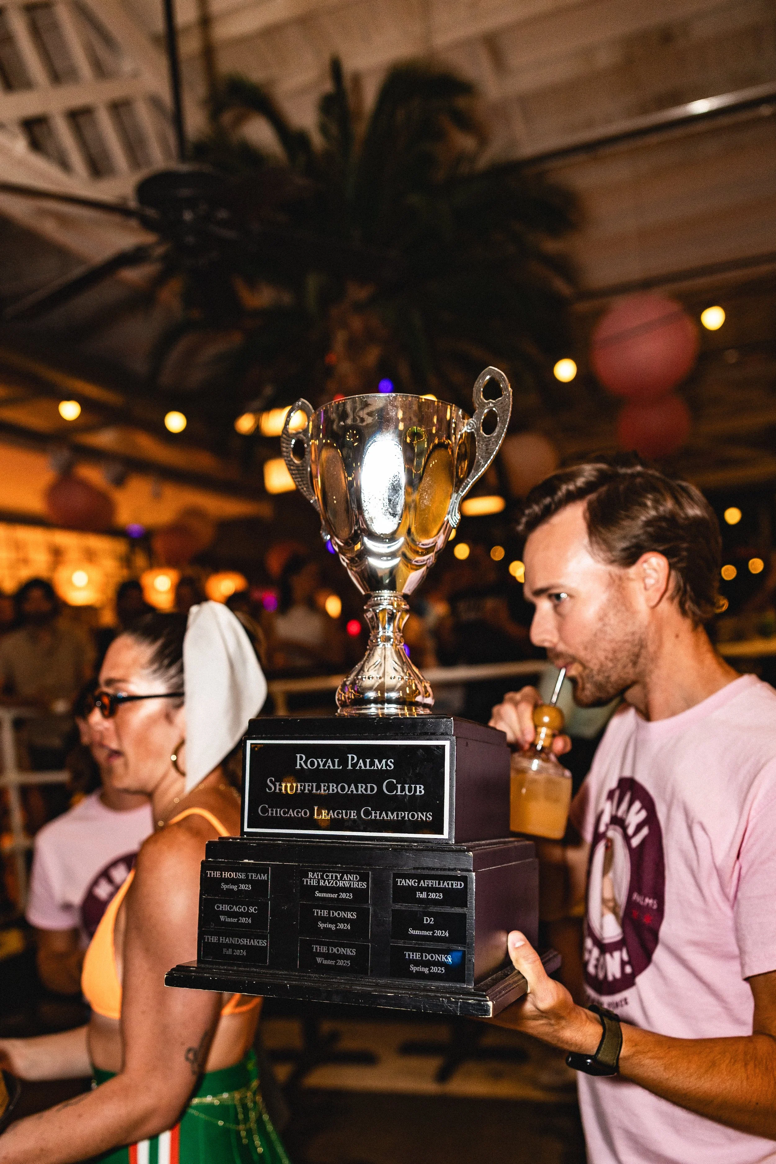 Man holding a trophy at a celebration event in a decorated indoor space, with a woman nearby and people in the background.