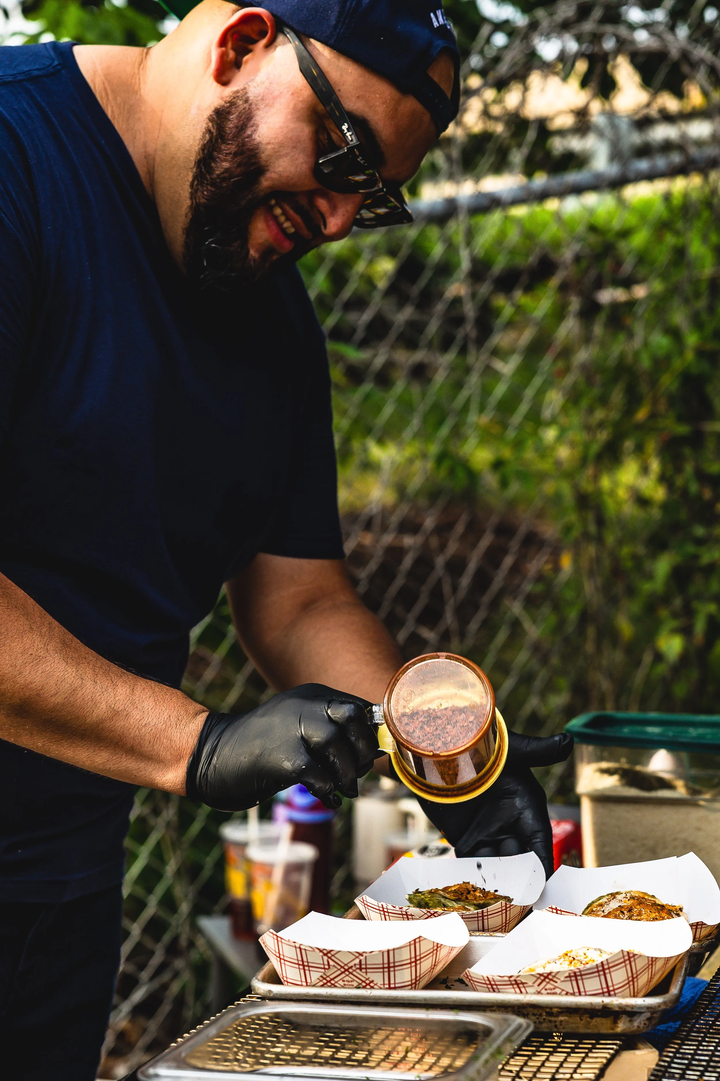 A man wearing a black shirt, black gloves, sunglasses, and a backwards baseball cap is pouring barbecue sauce on food in paper trays at an outdoor event.