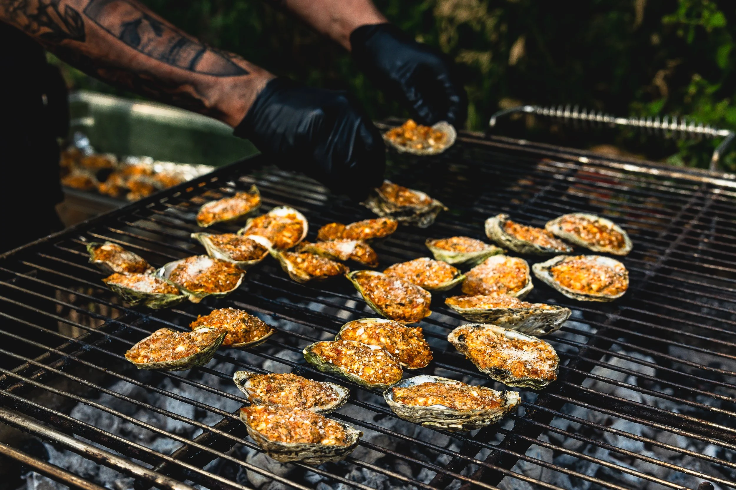 Person wearing black gloves grilling oysters with a spicy topping on a barbecue grill outdoors.