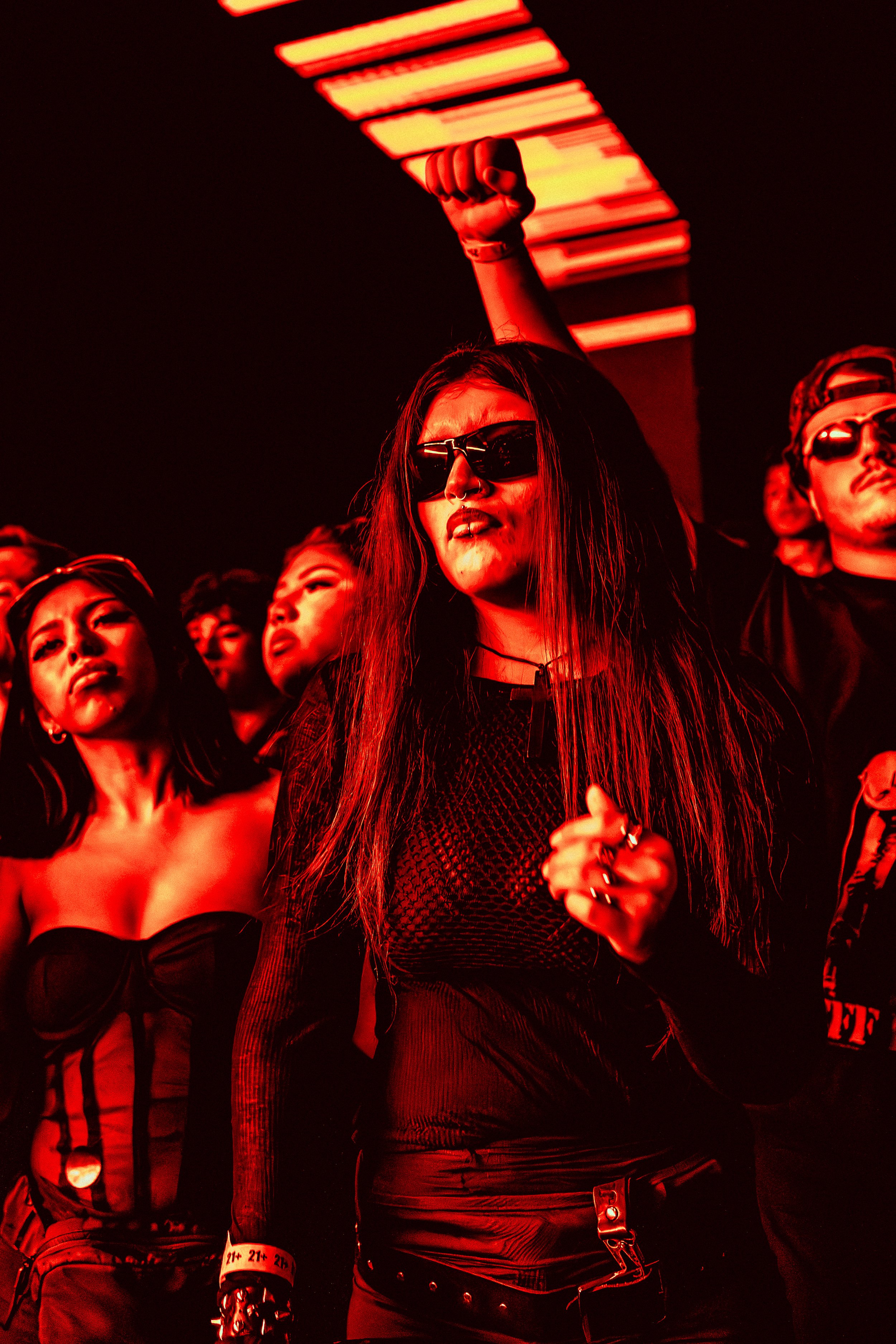 A group of young people at a concert or club, with a red and black lighting theme. One woman in the center wears sunglasses, a black mesh top, and has long hair, with her arm raised in a fist. Others around her are dressed in black, some wearing sung