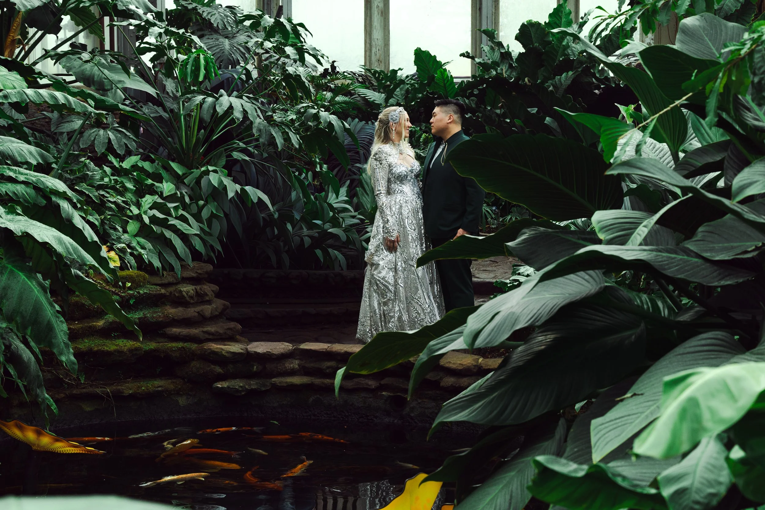 A couple in wedding attire standing close to each other amidst lush green tropical plants, with a pond and koi fish in the foreground.