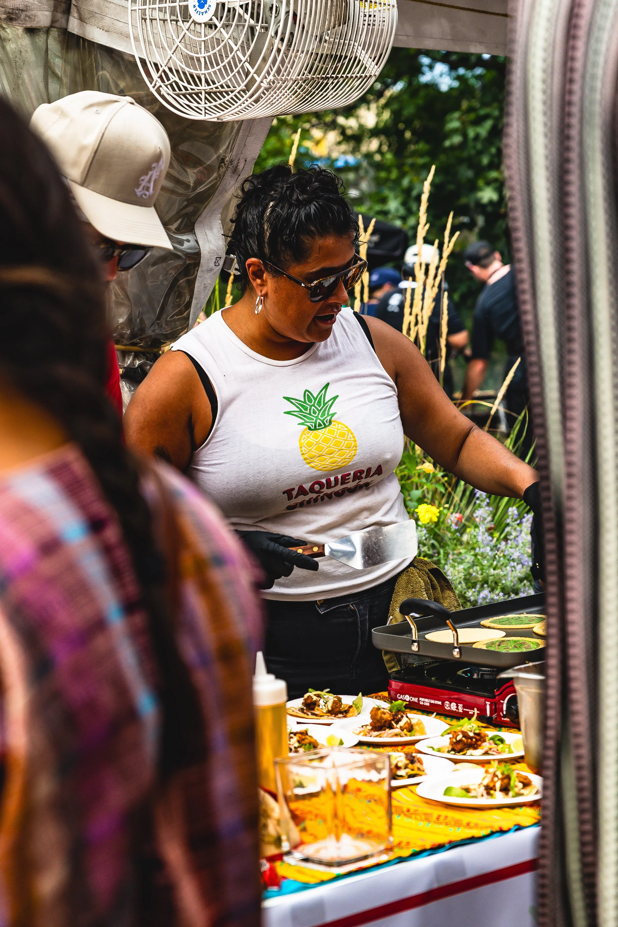 A woman wearing sunglasses and a white sleeveless shirt with a pineapple logo is cooking tacos at an outdoor event, with plates of prepared food on a table.