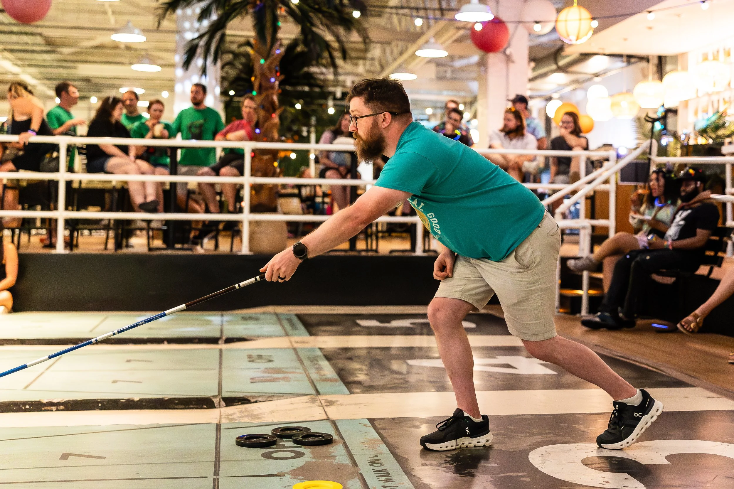 A man playing shuffleboard indoors, leaning forward to slide a disk on the game surface, with spectators watching from the background.