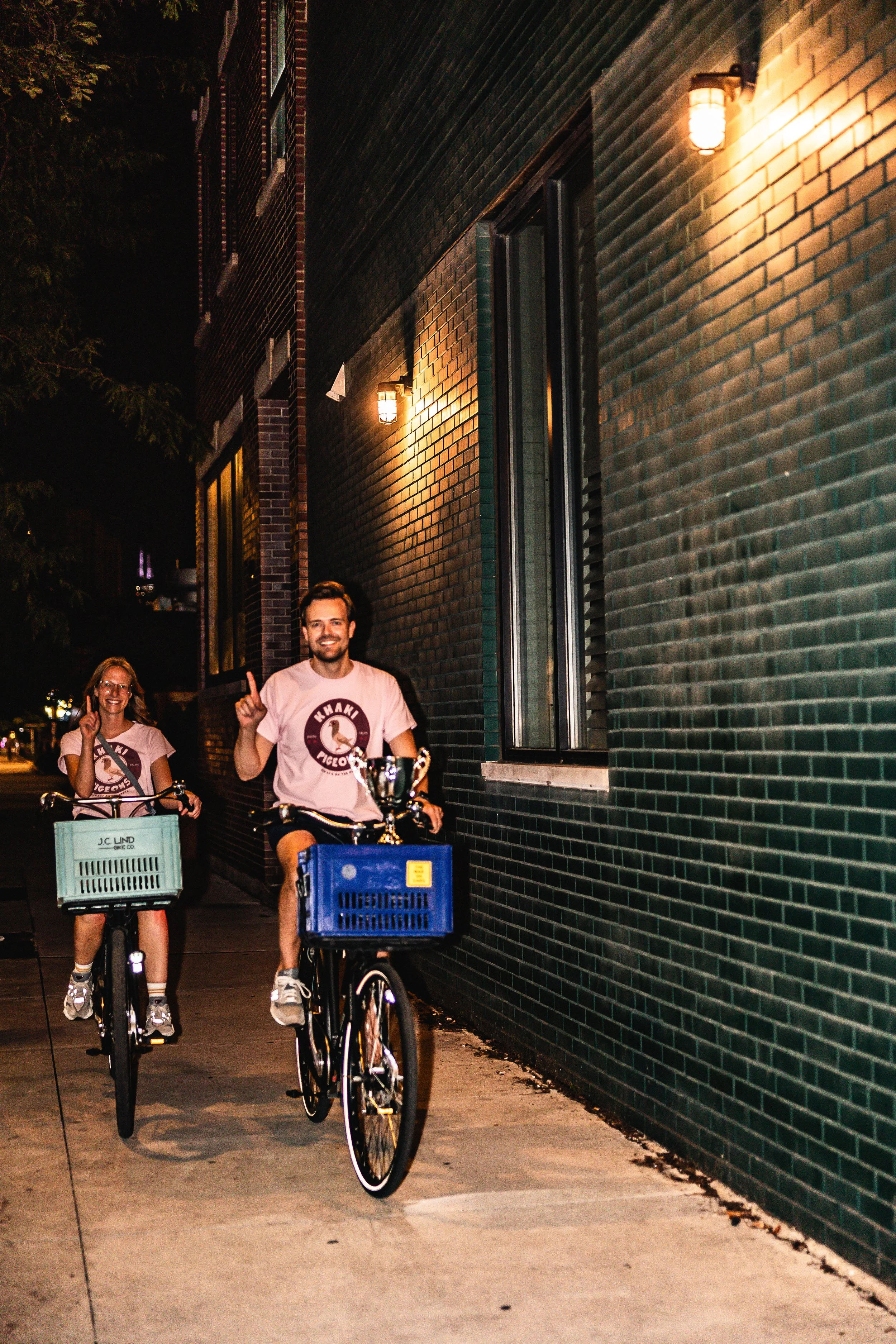 Two people riding bikes at night along a dark sidewalk next to a dark brick building, both wearing matching light-colored T-shirts, smiling and making a peace sign.