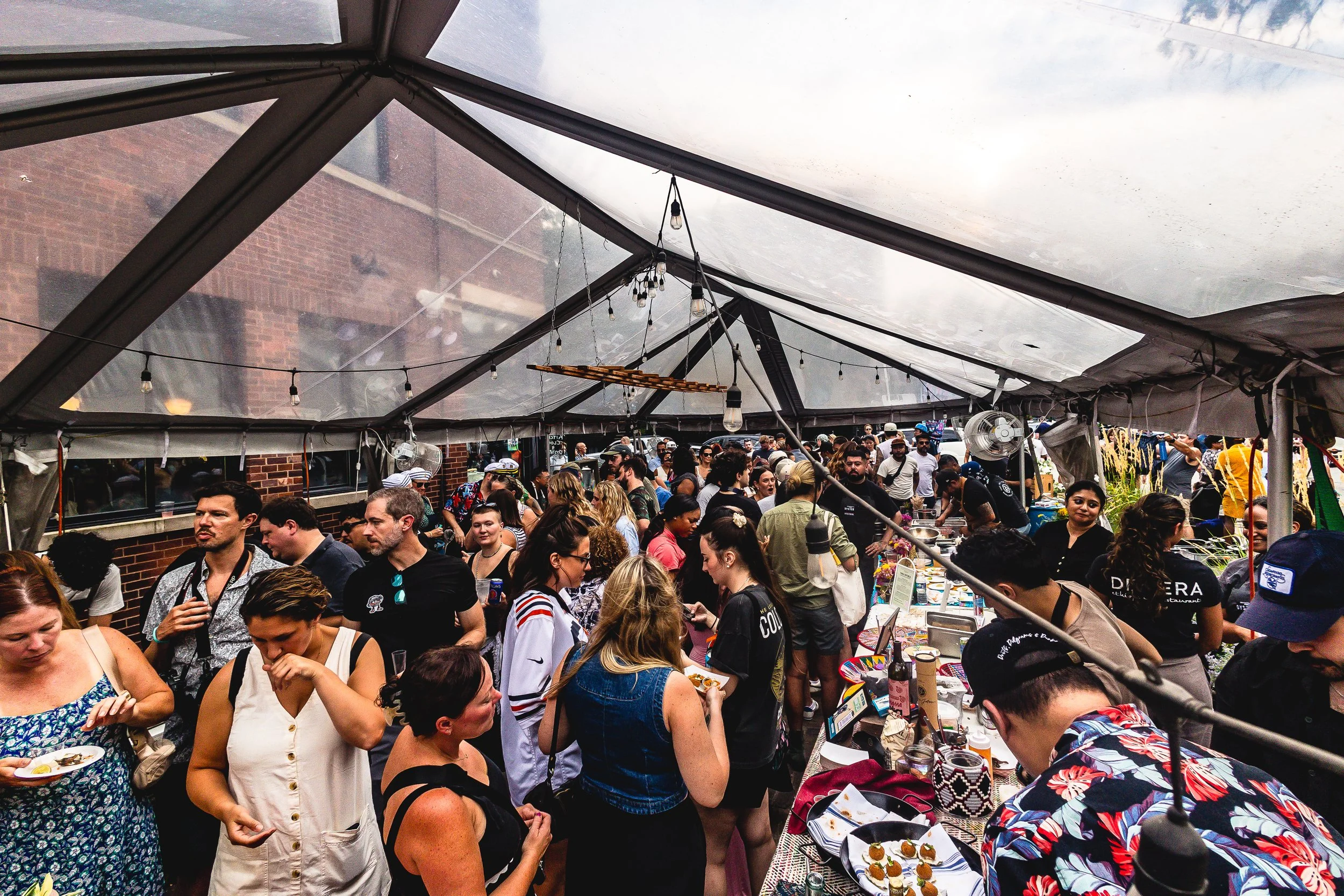 Crowd of people at an outdoor market or food festival under a tent with glass panels, with food stalls and lights