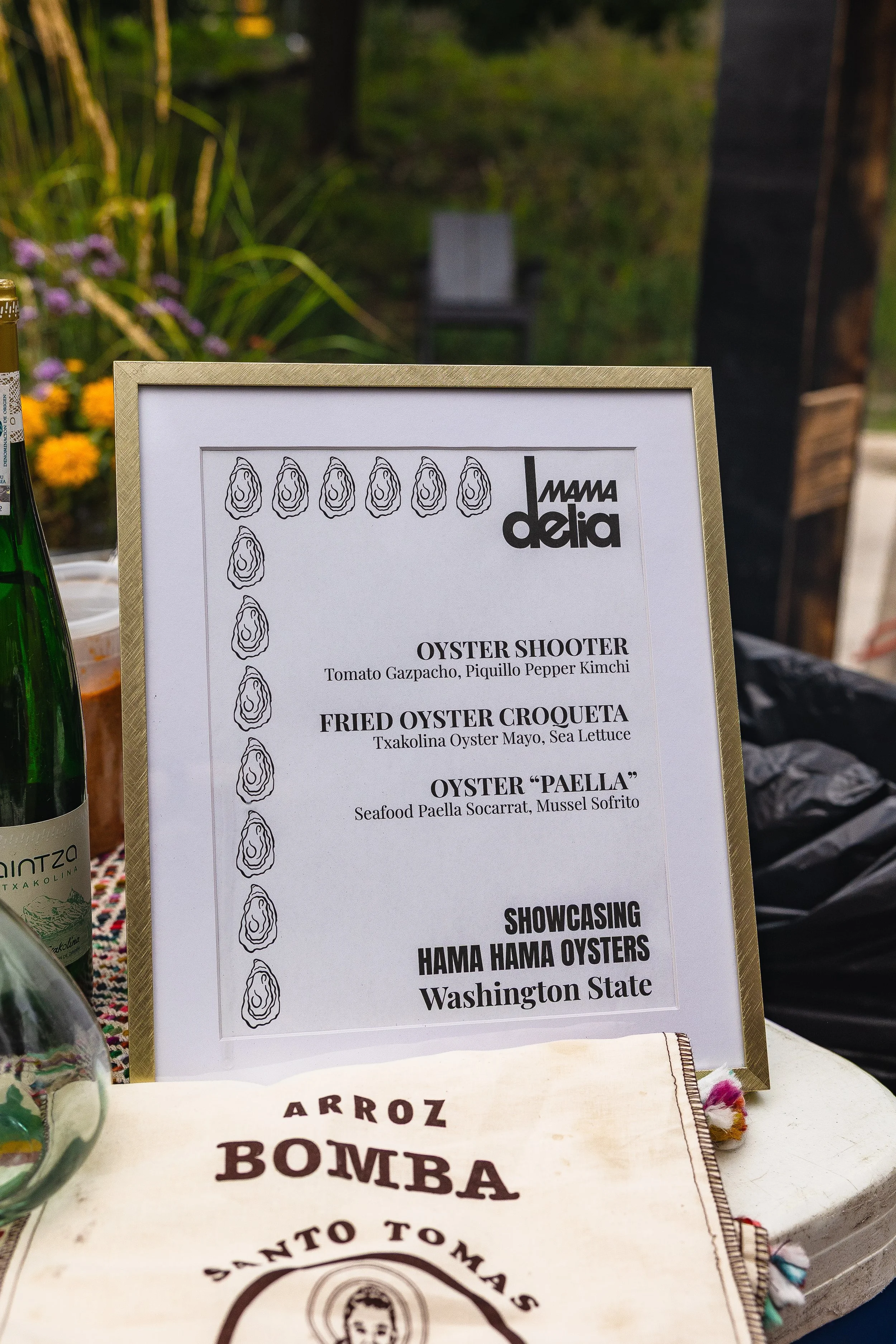 A menu stand displaying seafood dishes such as oyster shooter, fried oyster croqueta, and oyster paella at an outdoor event. The menu features illustrations of oyster shells along the border and promotes Hama Hama oysters from Washington State.