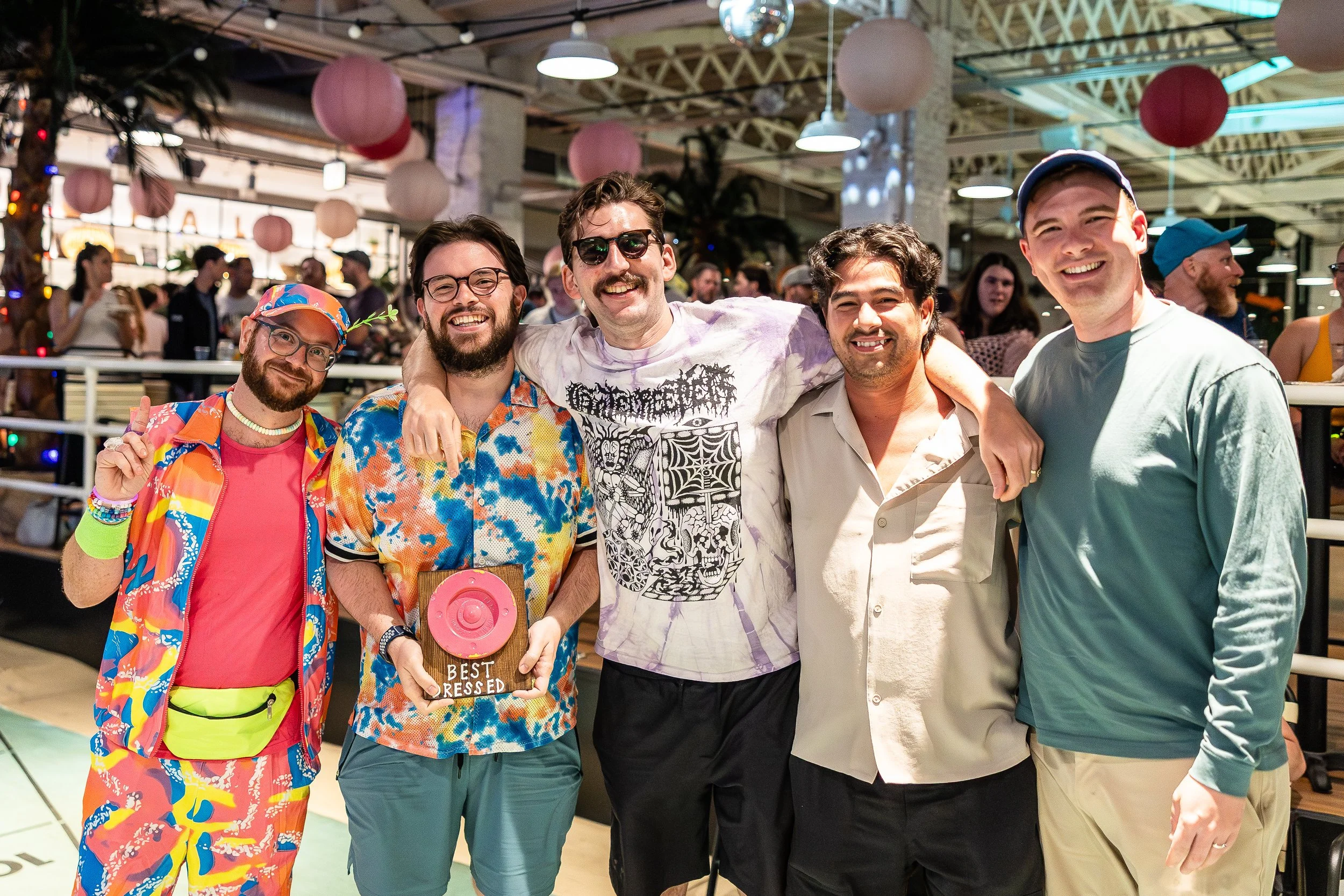 Group of five friends smiling and posing together at a lively indoor event, gathered closely with arms around each other in colorful casual attire.