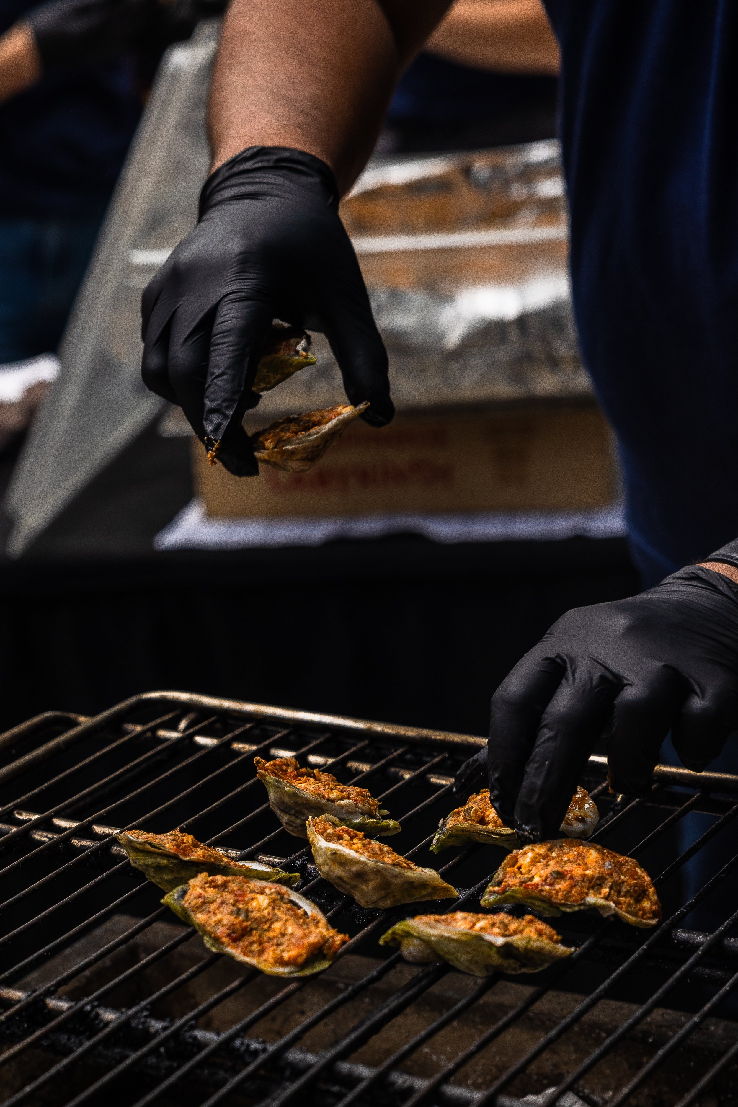 Person wearing black gloves placing stuffed oyster shells on a grill for cooking.