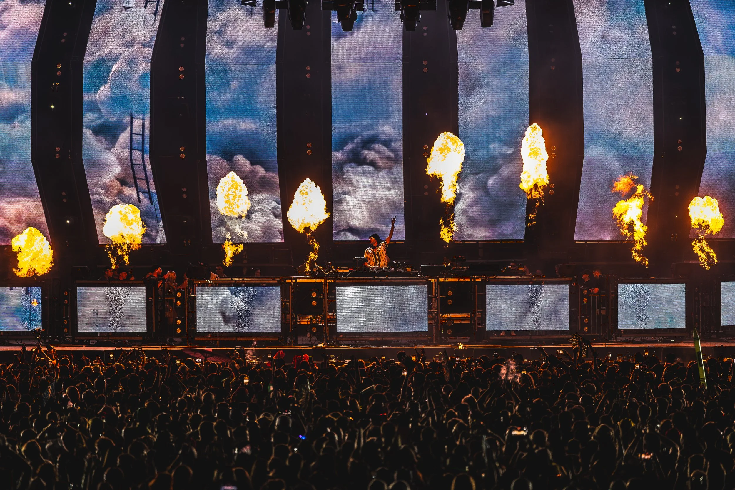 A DJ performs on stage at a concert with pyrotechnics and large screens displaying clouds in the background, while a large crowd watches in the foreground.