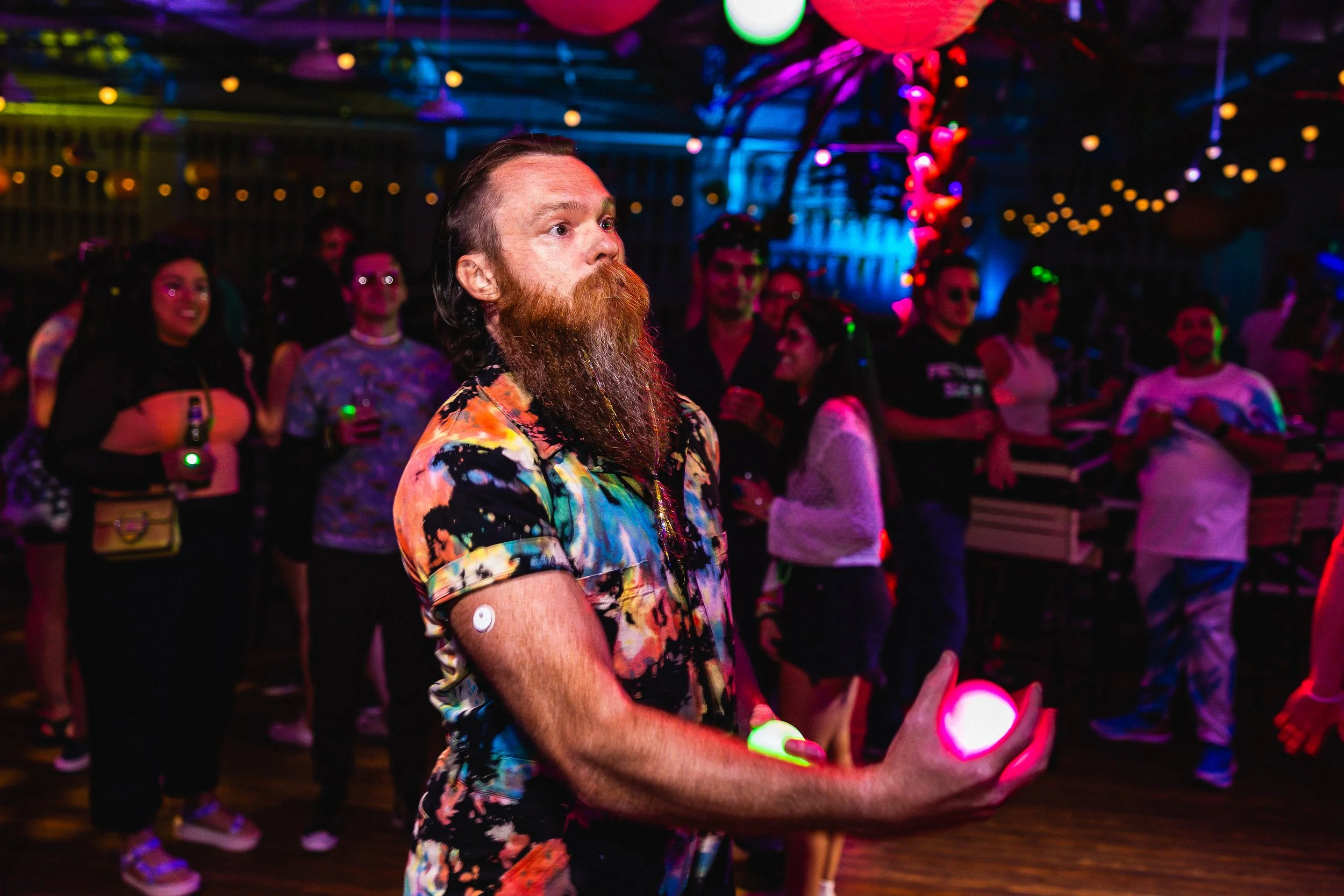 A man with a beard and mustache juggling glowing balls at a party with colorful lights, surrounded by people holding drinks and dancing.