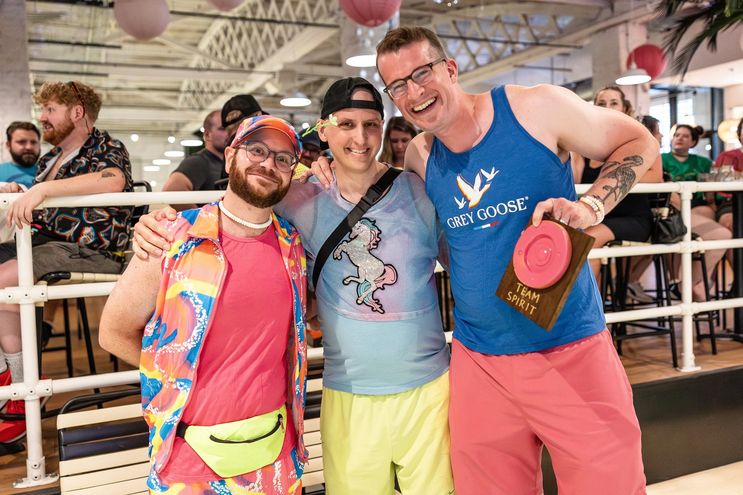 Three smiling men at an indoor event, one holding a pink disc with a sign that reads 'Team Spirit,' surrounded by seated and standing people.