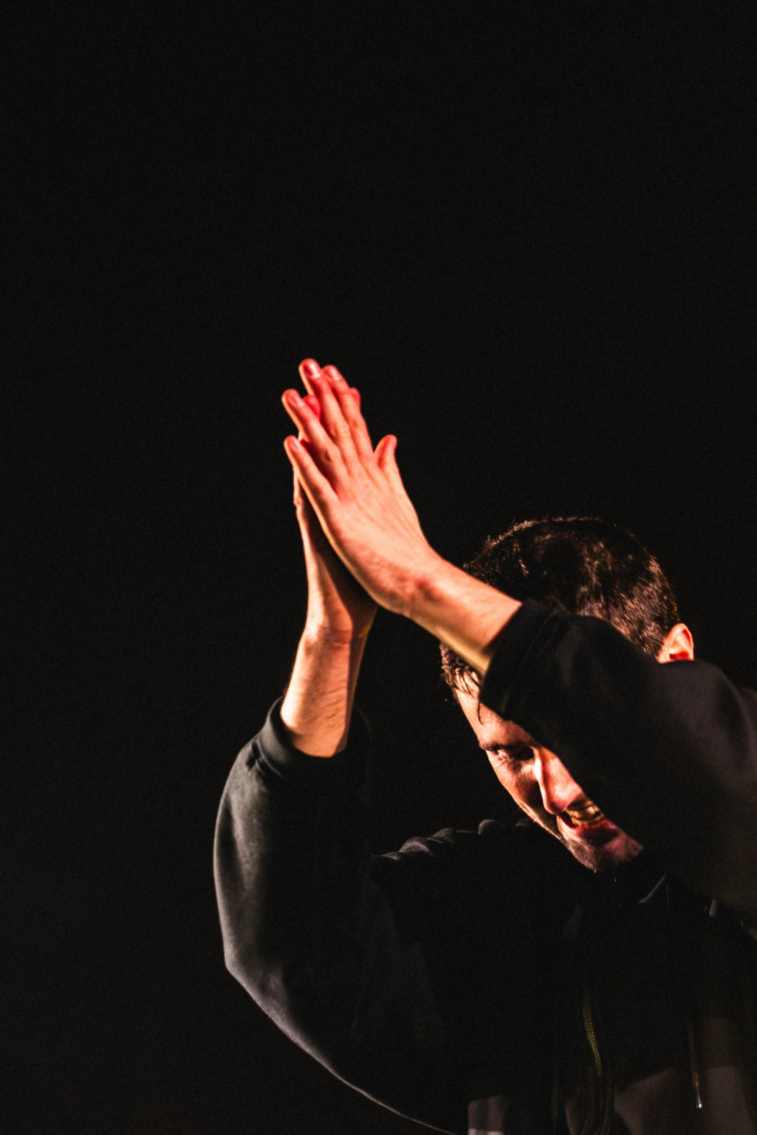 Fred Again with dark hair, wearing a black jacket, is smiling and clapping his hands together with a dark background.