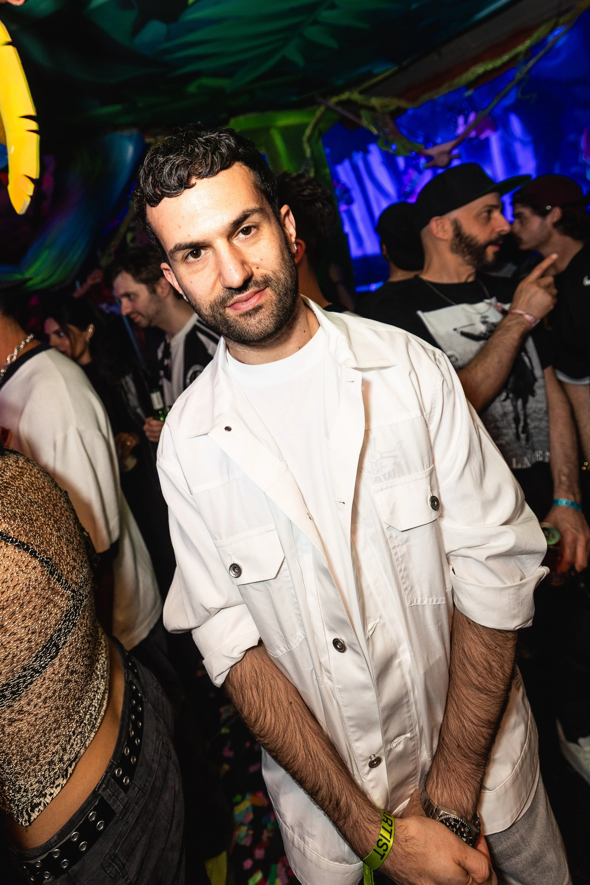 A man with dark hair, beard, and earrings wearing a white jacket and white t-shirt at a crowded party with colorful lighting.