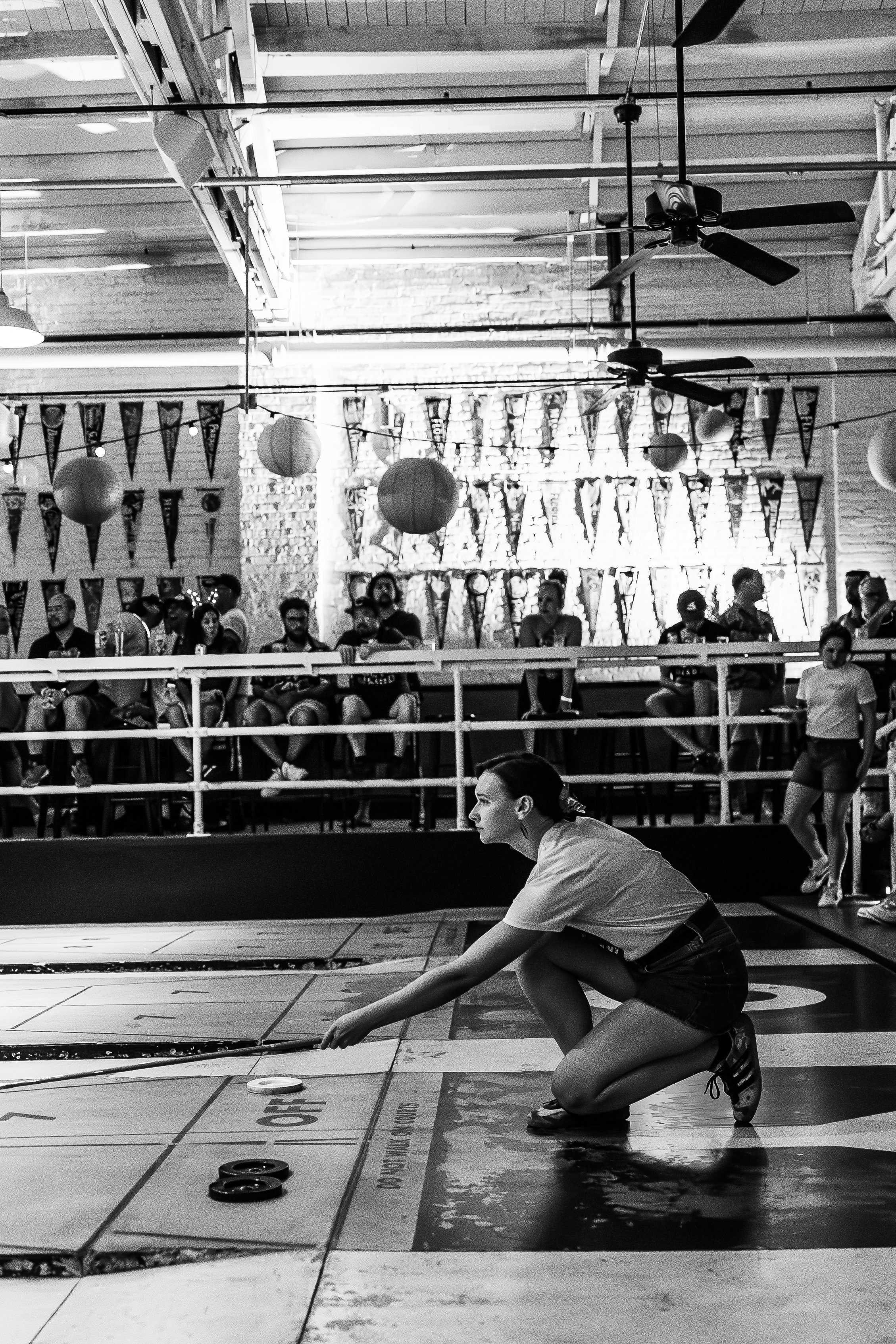 A woman crouching on the floor in a competitive indoor curling game, with spectators seated in the background and decorations hanging in the venue.