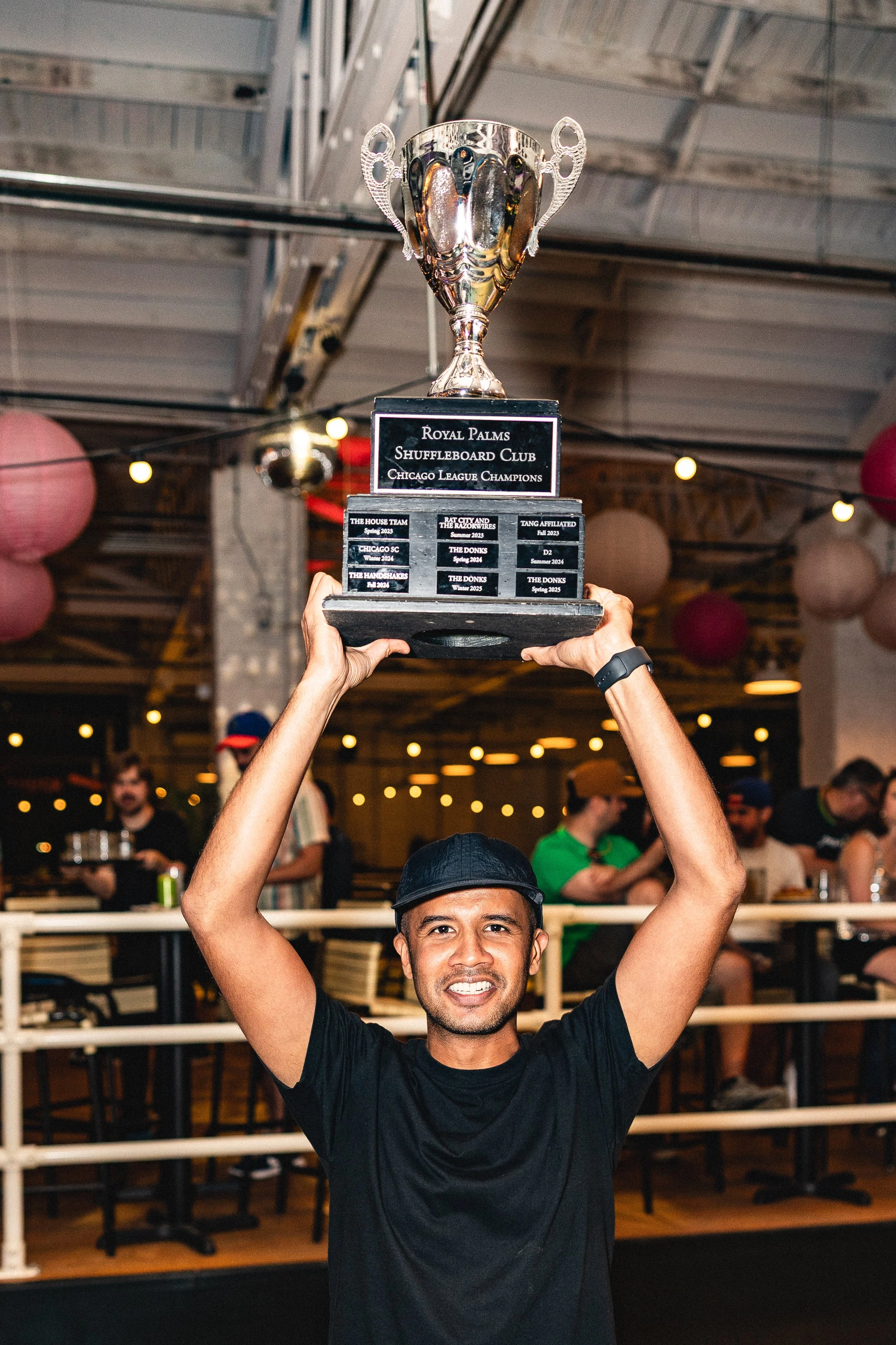 A man holding a trophy above his head in a celebration setting, with other people in the background, and red and pink paper lanterns hanging from the ceiling.