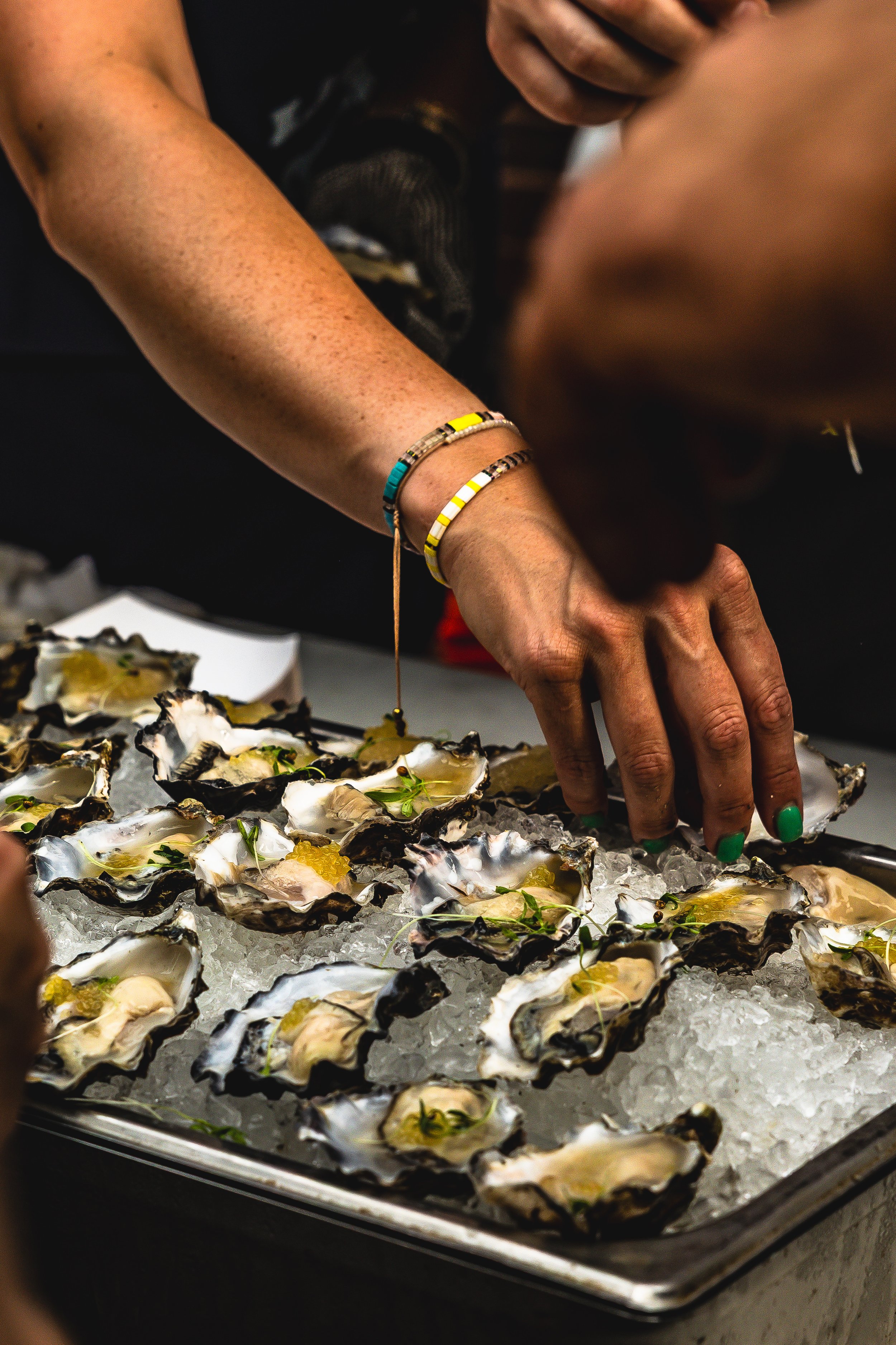 Close-up of a person with green-painted nails picking up fresh oysters on a bed of ice, garnished with herbs, at a seafood restaurant or market.