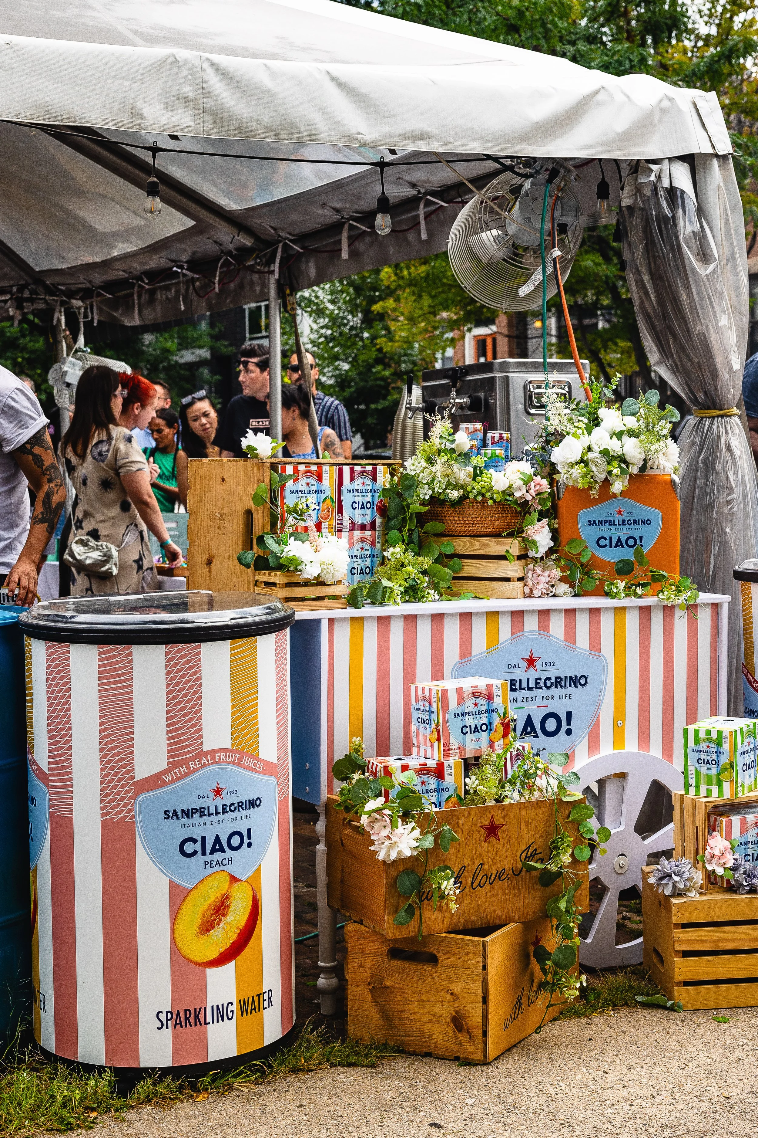 A fruit juice stand decorated with flowers and banners with people in line in the background at an outdoor event.