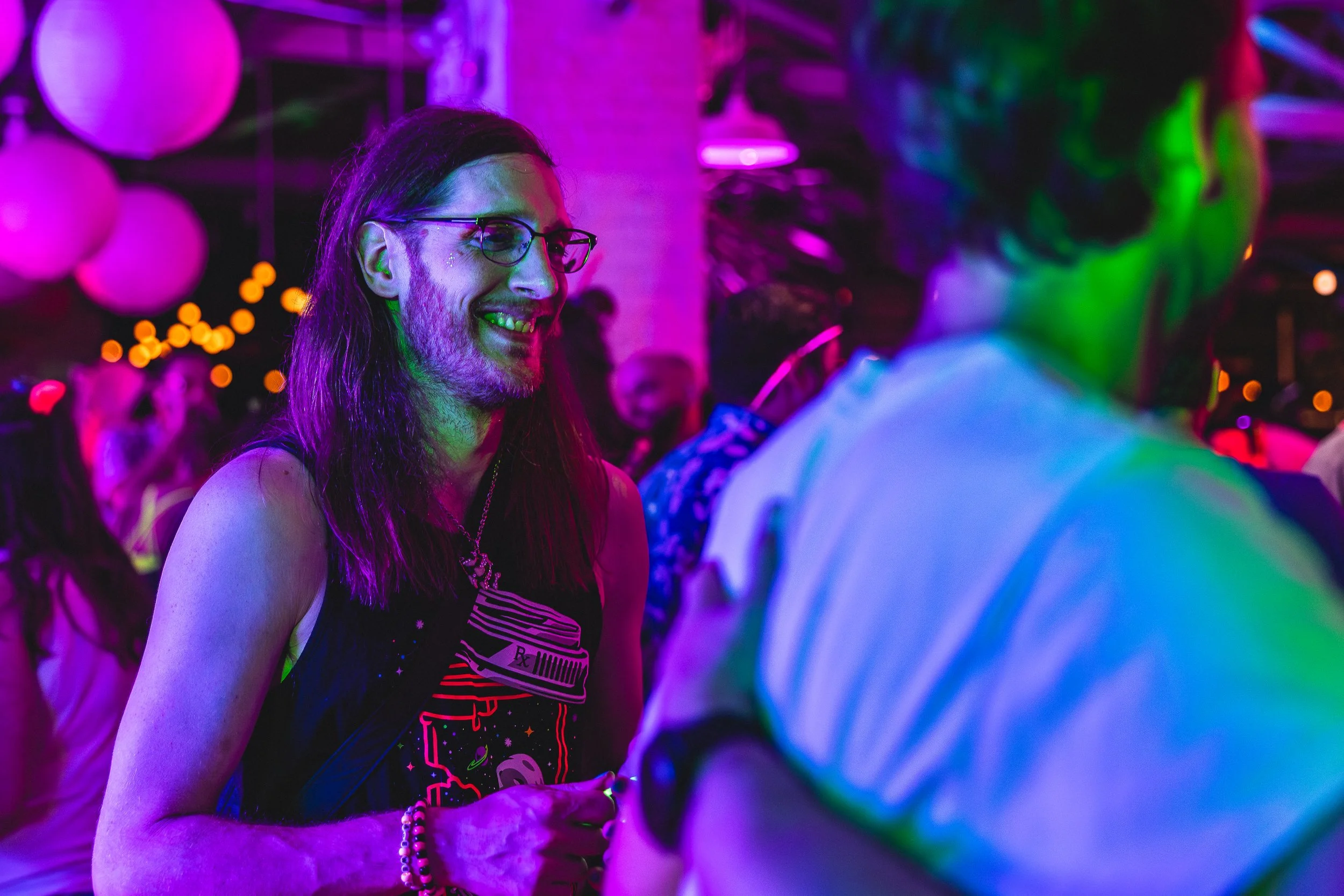 Two people smiling and talking at a party with colorful neon lights, with one person wearing glasses and a black shirt with a graphic design.