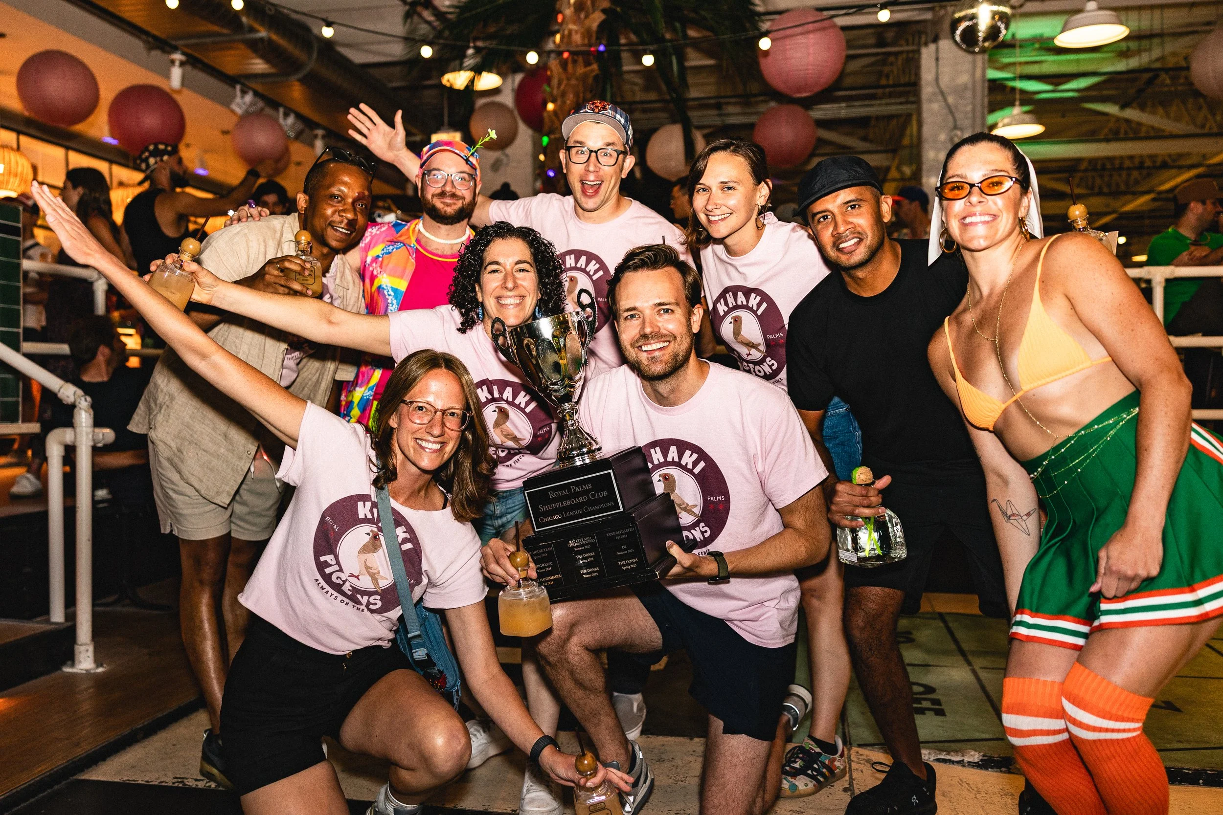A diverse group of people celebrating together at an indoor party, holding a trophy, with some wearing matching 'Khaki Pugys' T-shirts, in a lively venue decorated with paper lanterns and string lights.