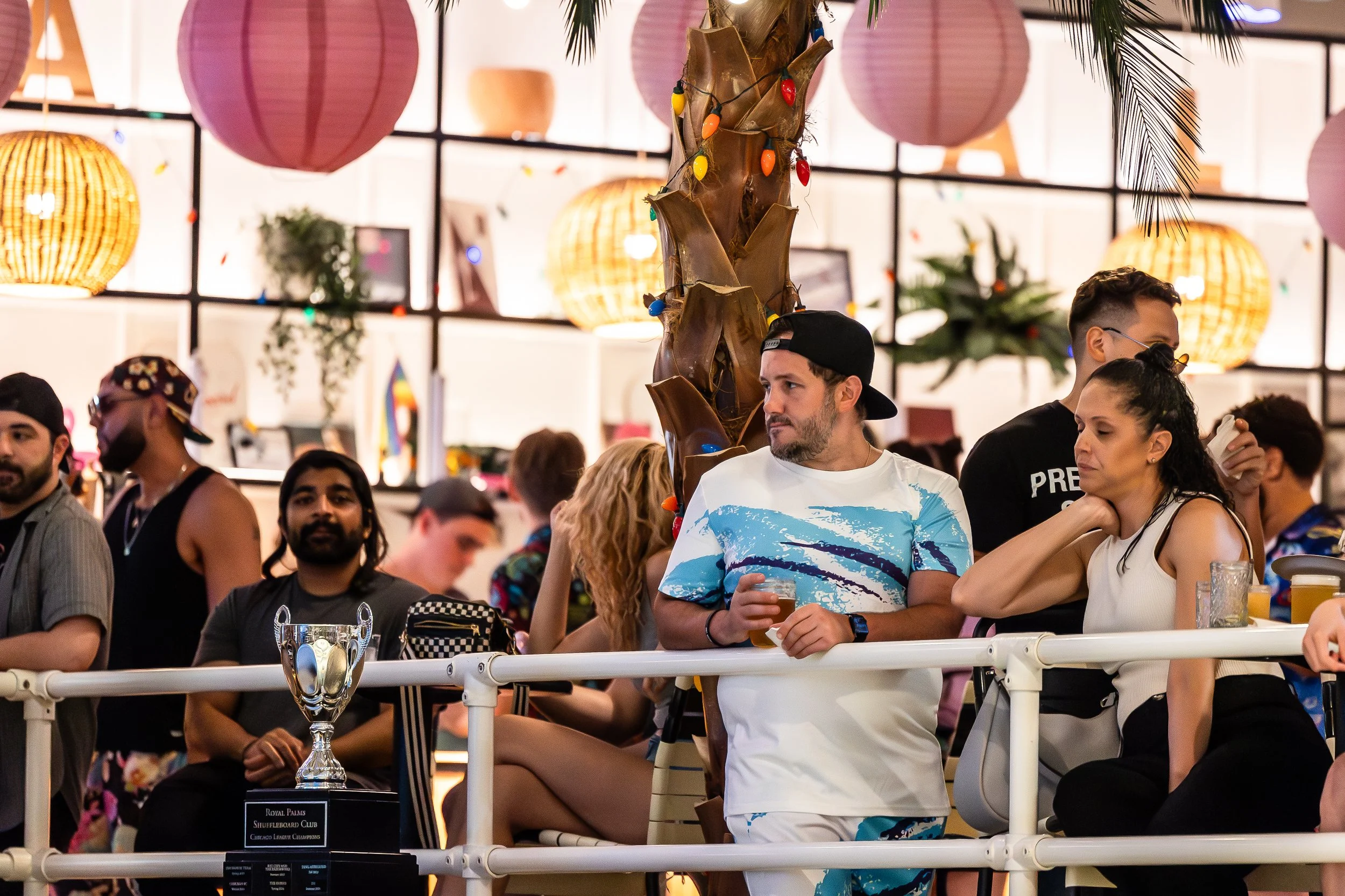 People gathered at a celebration or event with tropical decor, including hanging paper lanterns, a palm tree wrapped with colorful lights, and various decorations in the background.